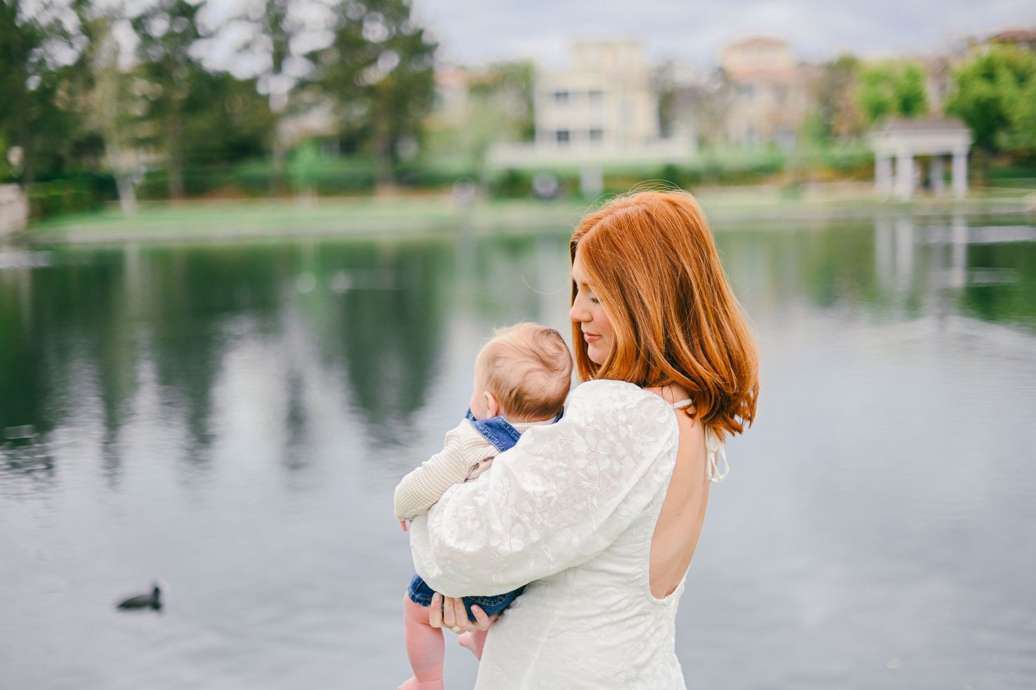 Woman in white dress holds baby while standing by peaceful lake at sunset.