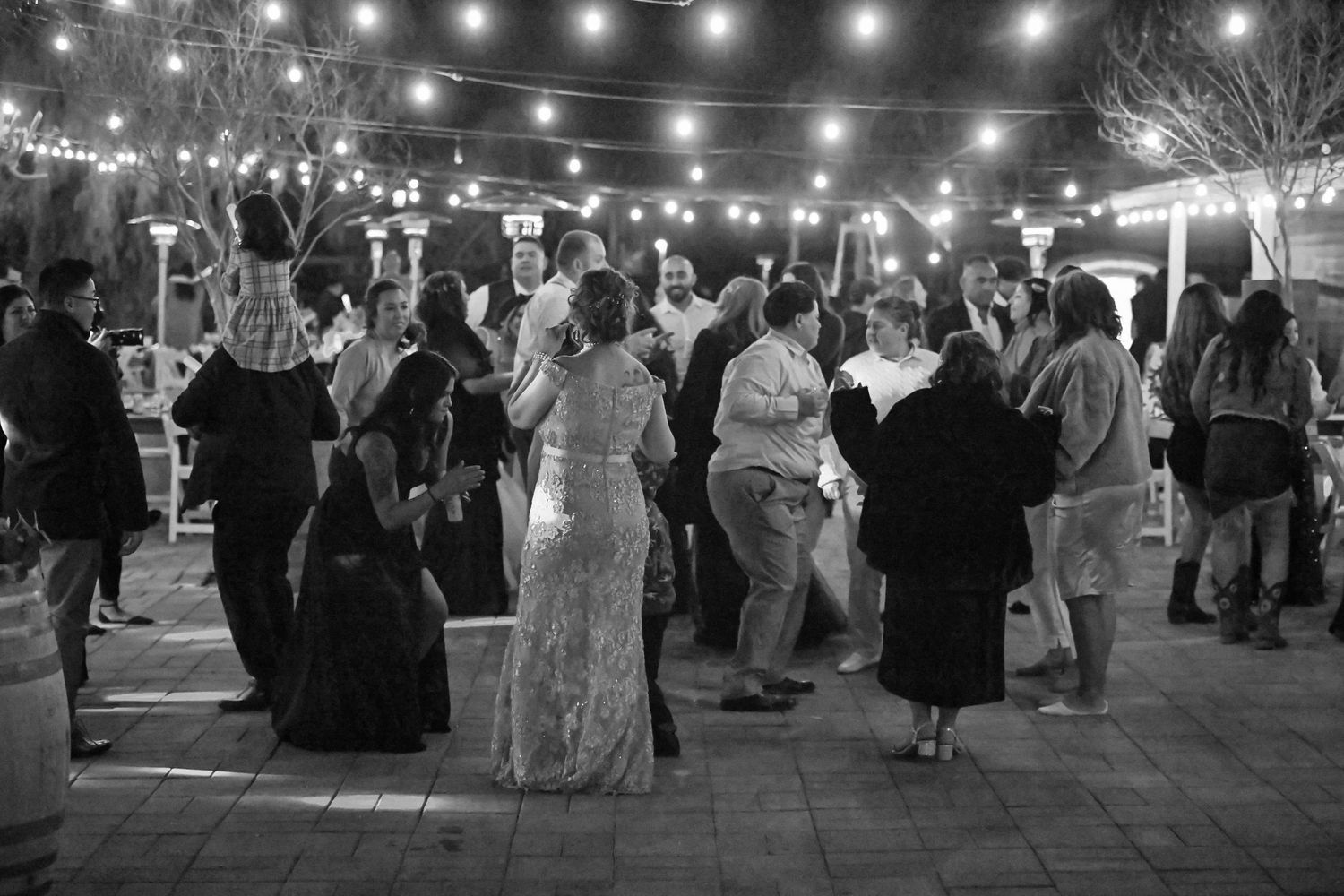Wedding guests dance under twinkling string lights at an outdoor evening reception.