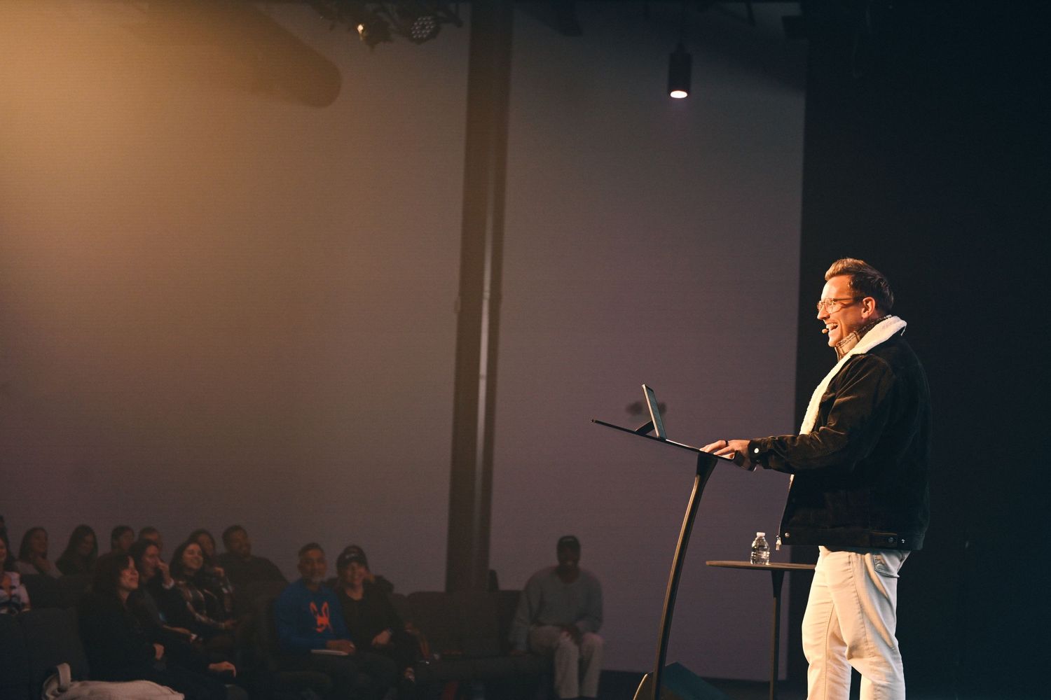A presenter speaks at a podium under bright stage lighting during an indoor event.