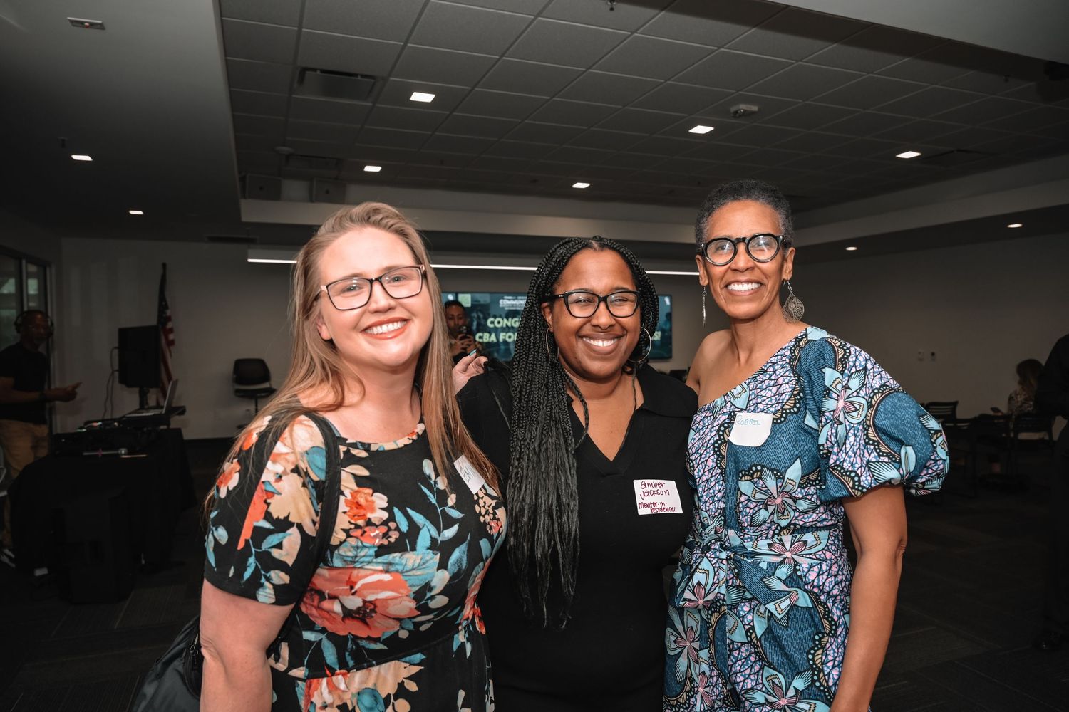 Three colleagues wearing colorful patterned outfits smile together at a professional event or meeting.