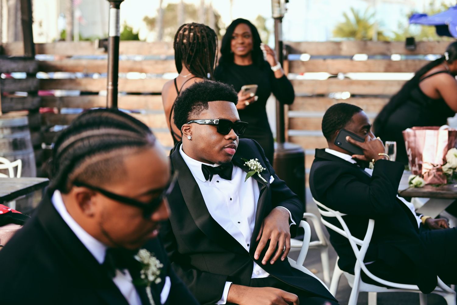 A group of well-dressed people in formal black tuxedos and sunglasses sitting outdoors at an event gathering.