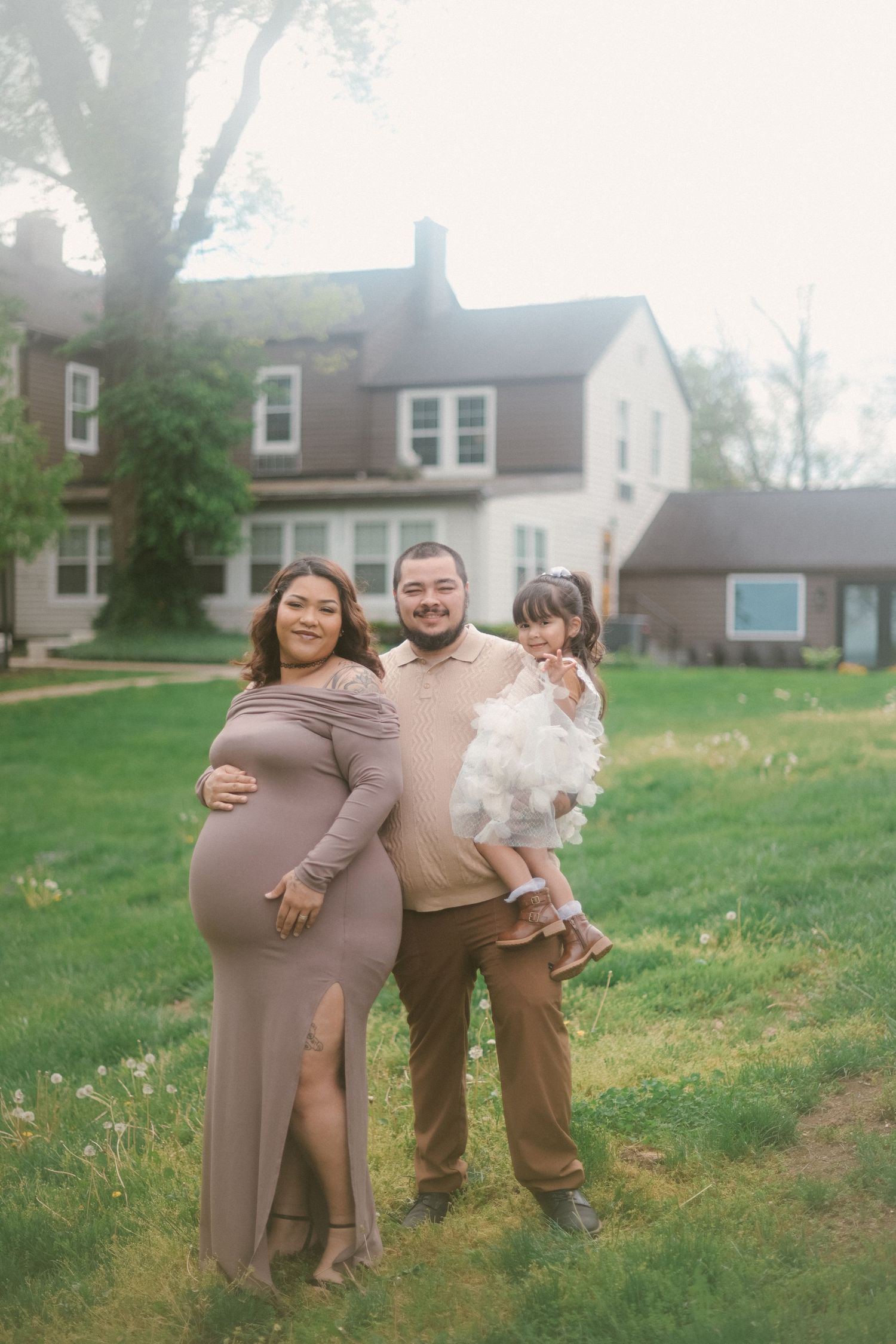 Family poses together on lawn in front of traditional house during maternity photoshoot.