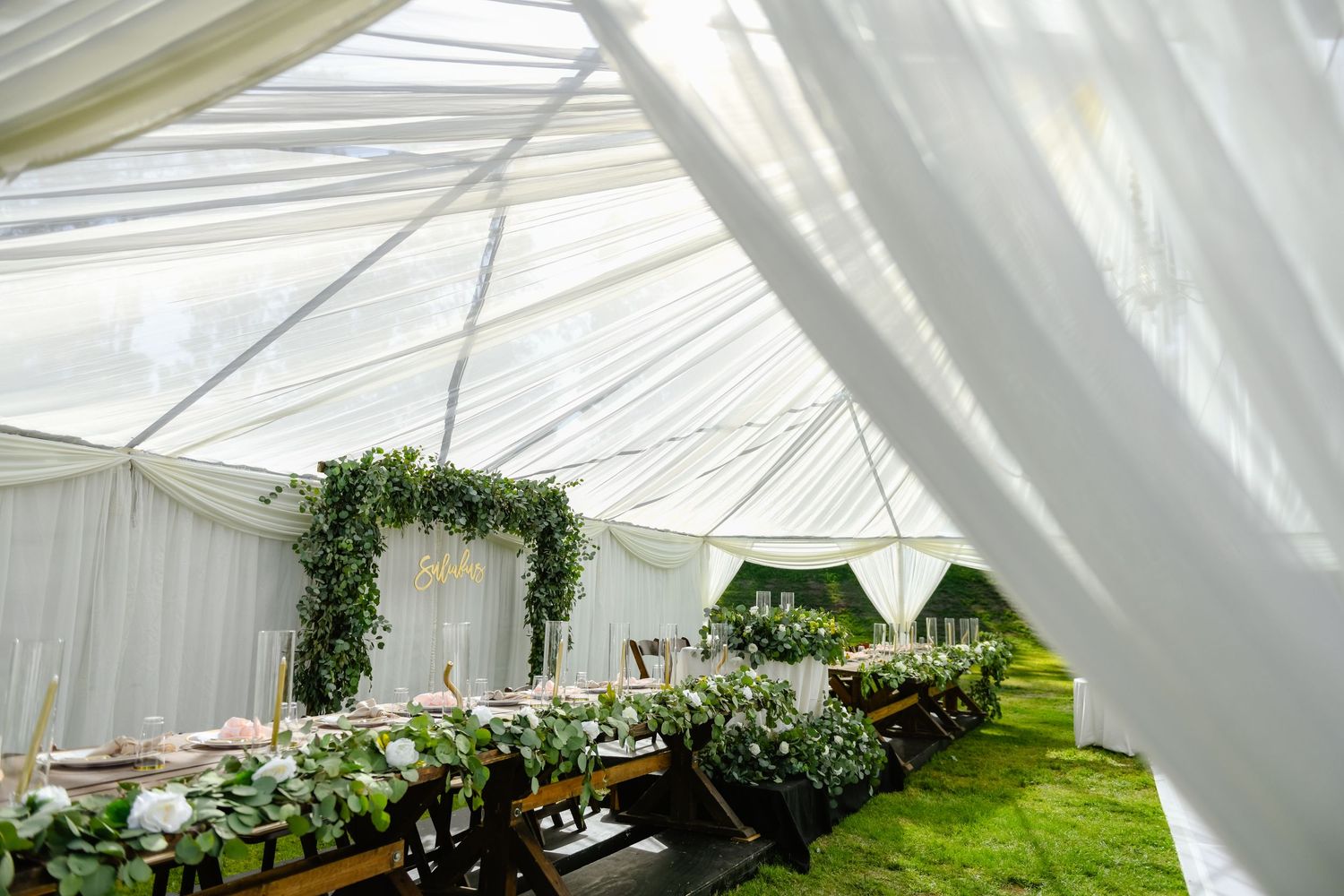 White wedding tent interior with elegant greenery garland arch and rustic table settings for a garden reception.