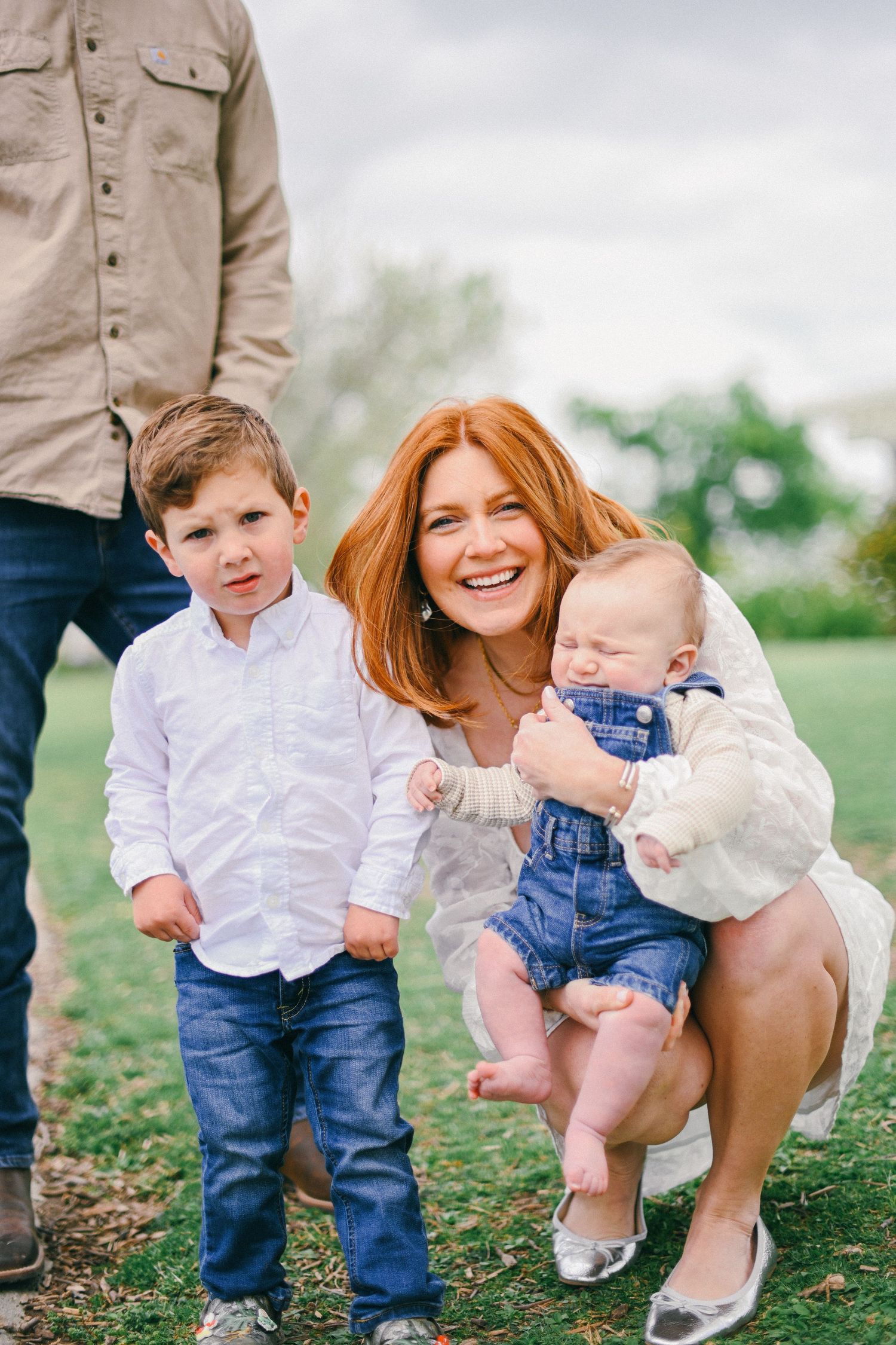 Family portrait outdoors with mom in white holding baby while son stands nearby.