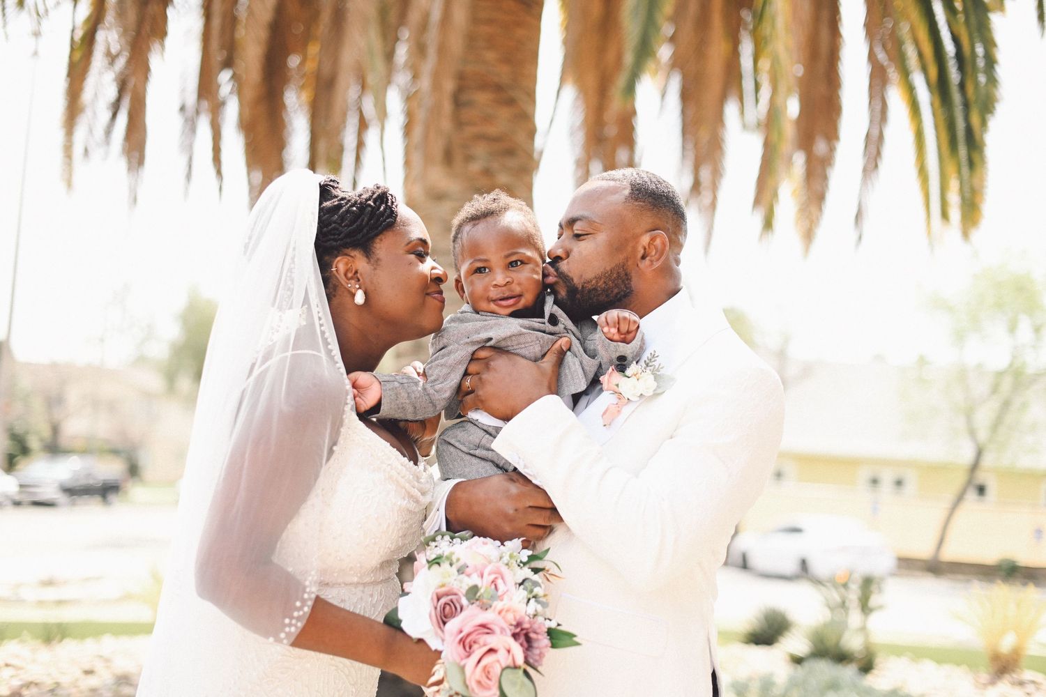 Wedding couple kisses their baby under palm trees in outdoor setting.