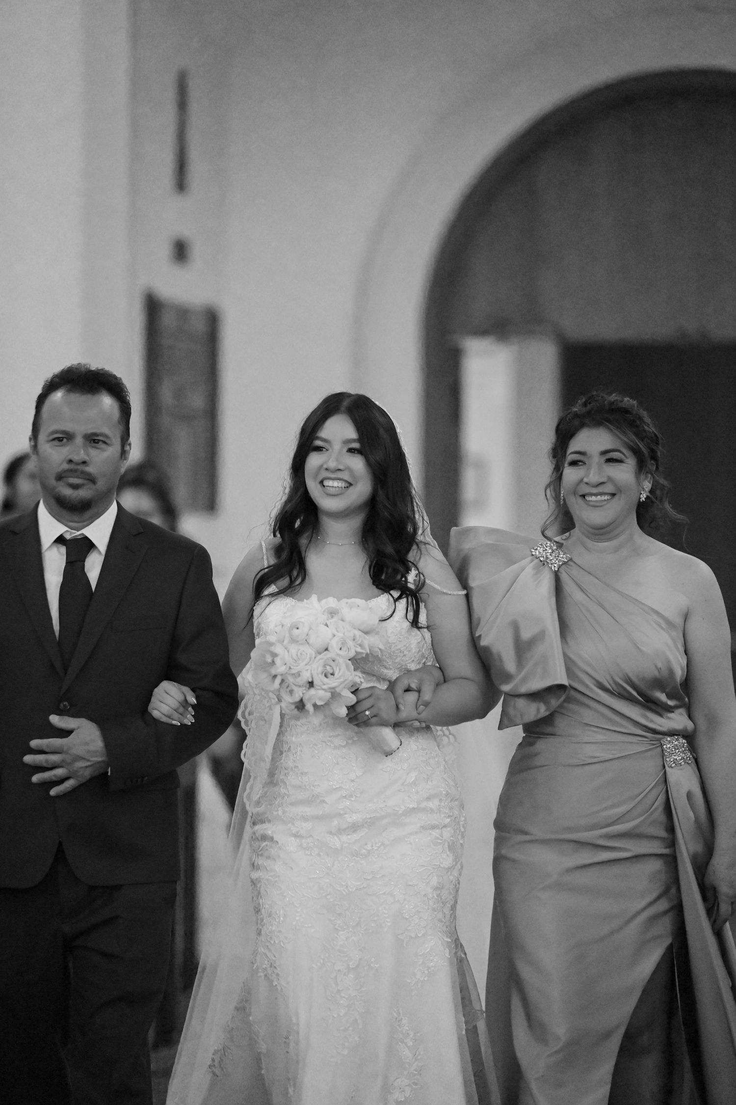 Bride in white lace gown walks down aisle with two attendants in formal attire.