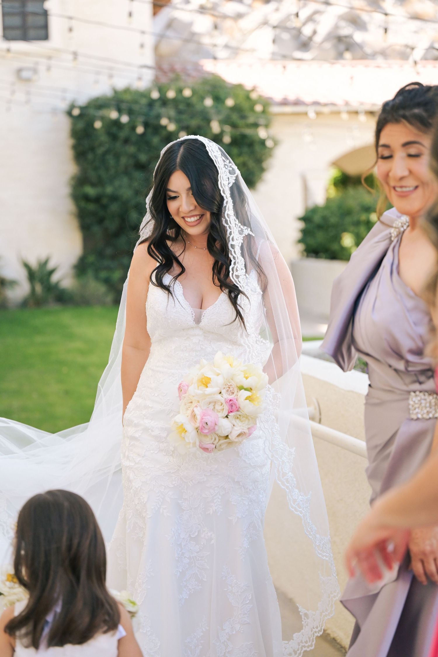 Bride in lace dress and long veil smiles while holding pastel bouquet during outdoor ceremony.