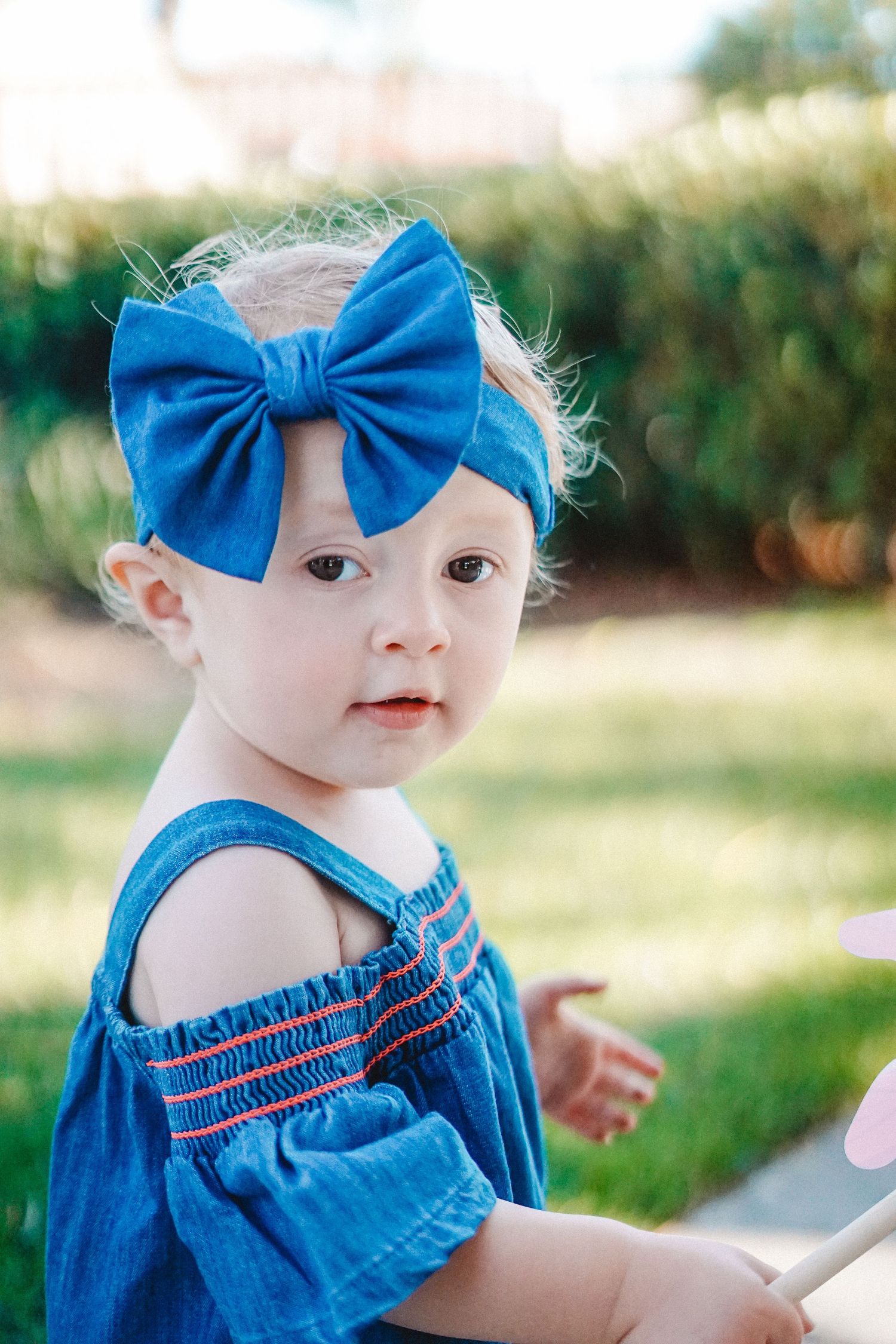 Baby wearing blue dress and matching bow headband sits outdoors in natural lighting.