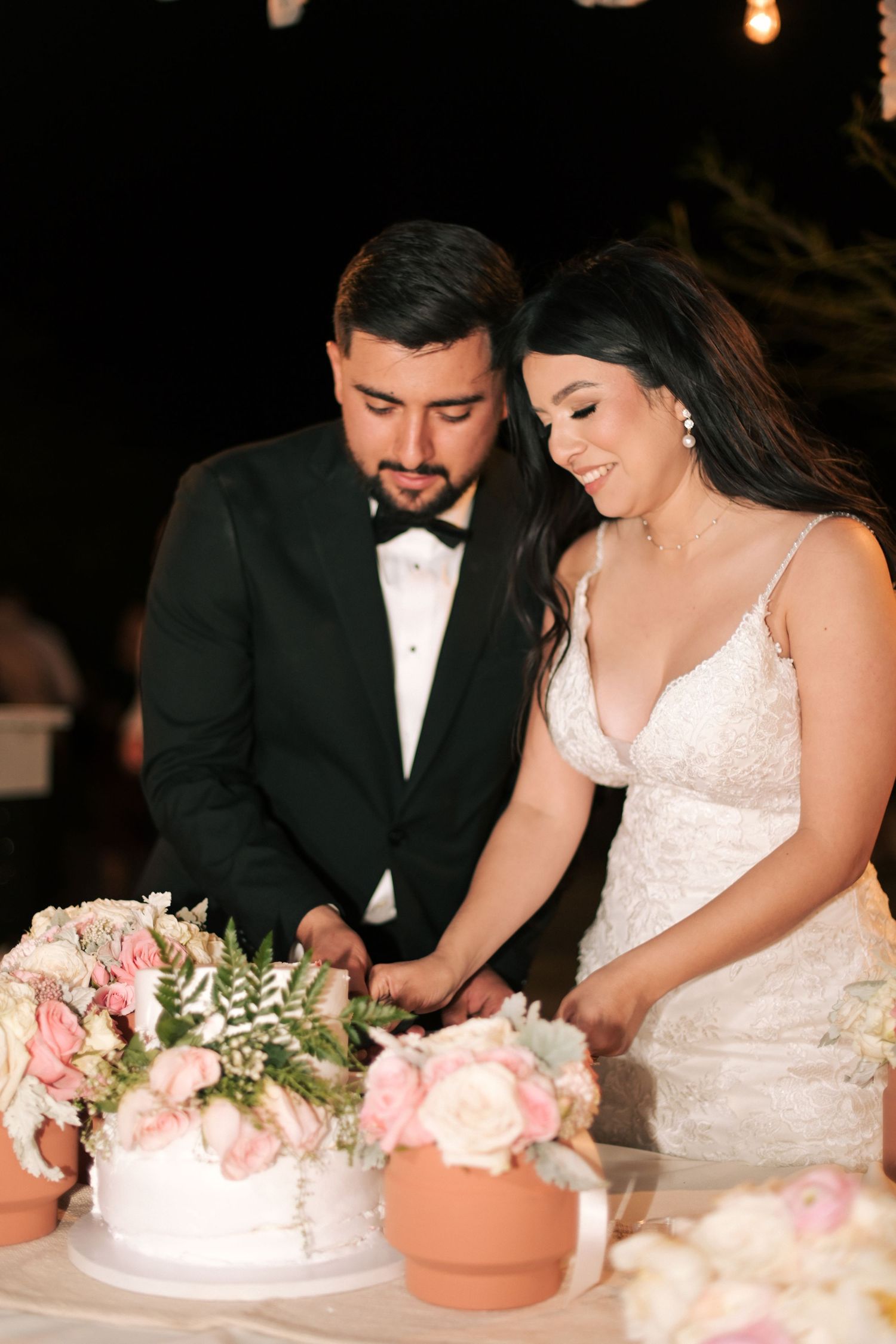 Newlyweds cut their wedding cake together surrounded by floral arrangements at evening reception.