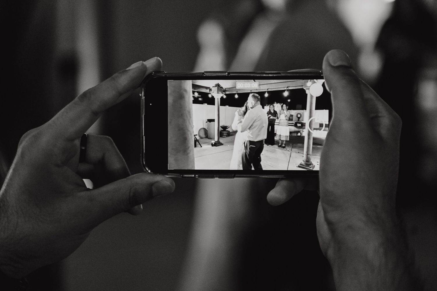 Hands hold a smartphone capturing wedding moments in black and white photography.