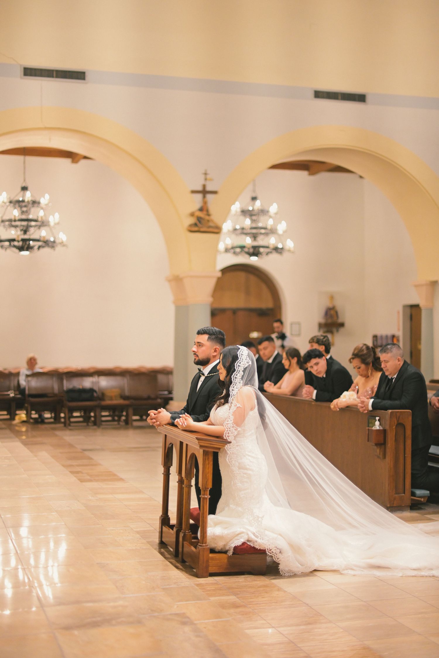 A couple kneels at the altar during their wedding ceremony in a grand church with arched ceilings.