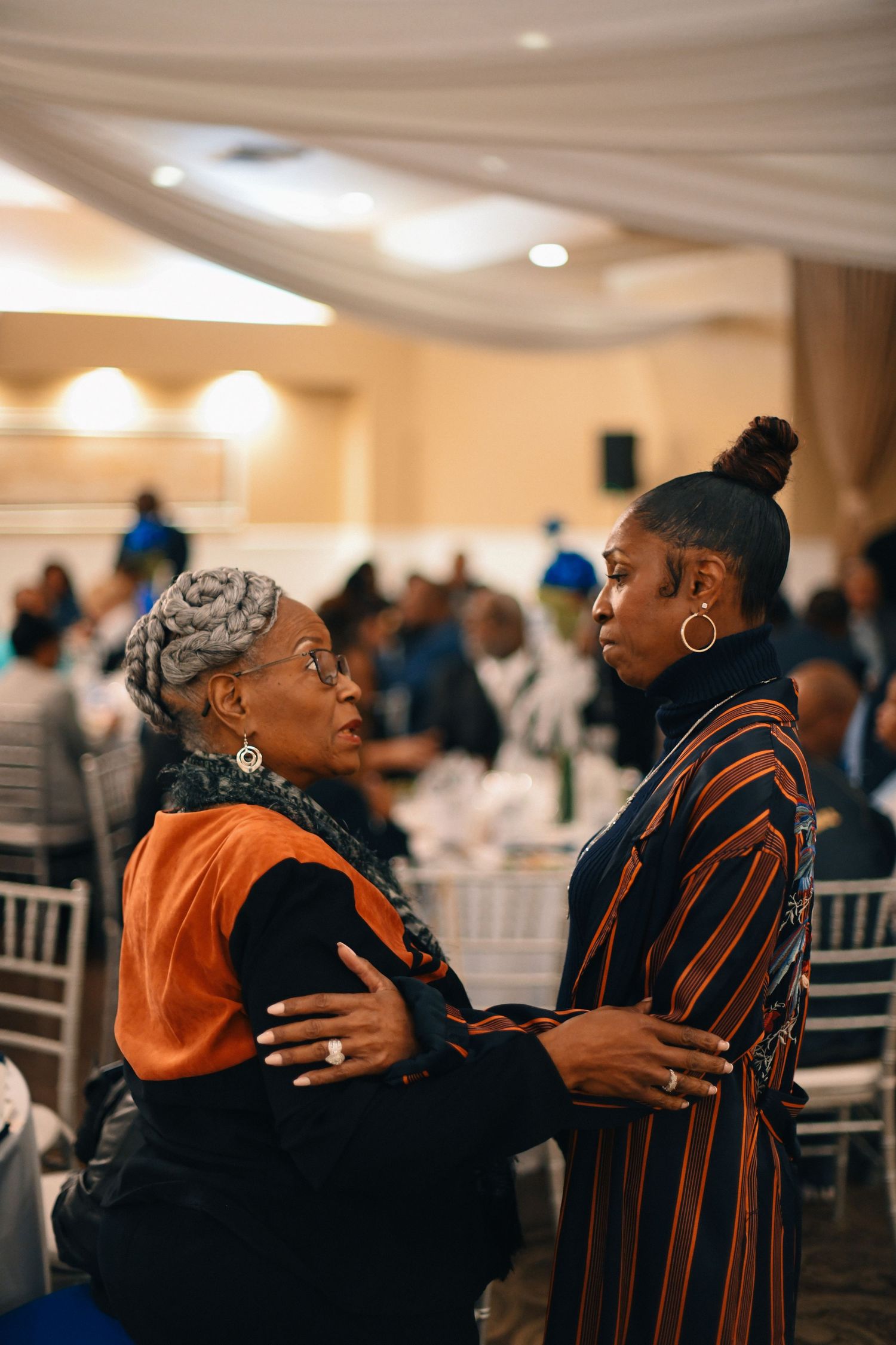 Two people share an intimate moment of connection during a formal event in an elegantly decorated banquet hall.