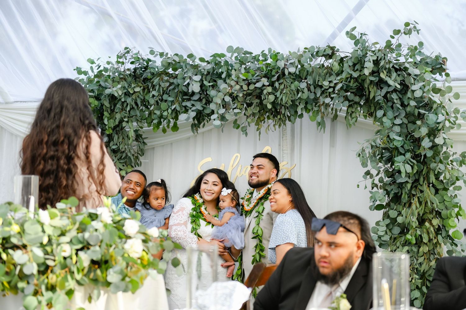 Celebration under a floral arch with greenery in a white tent setting.