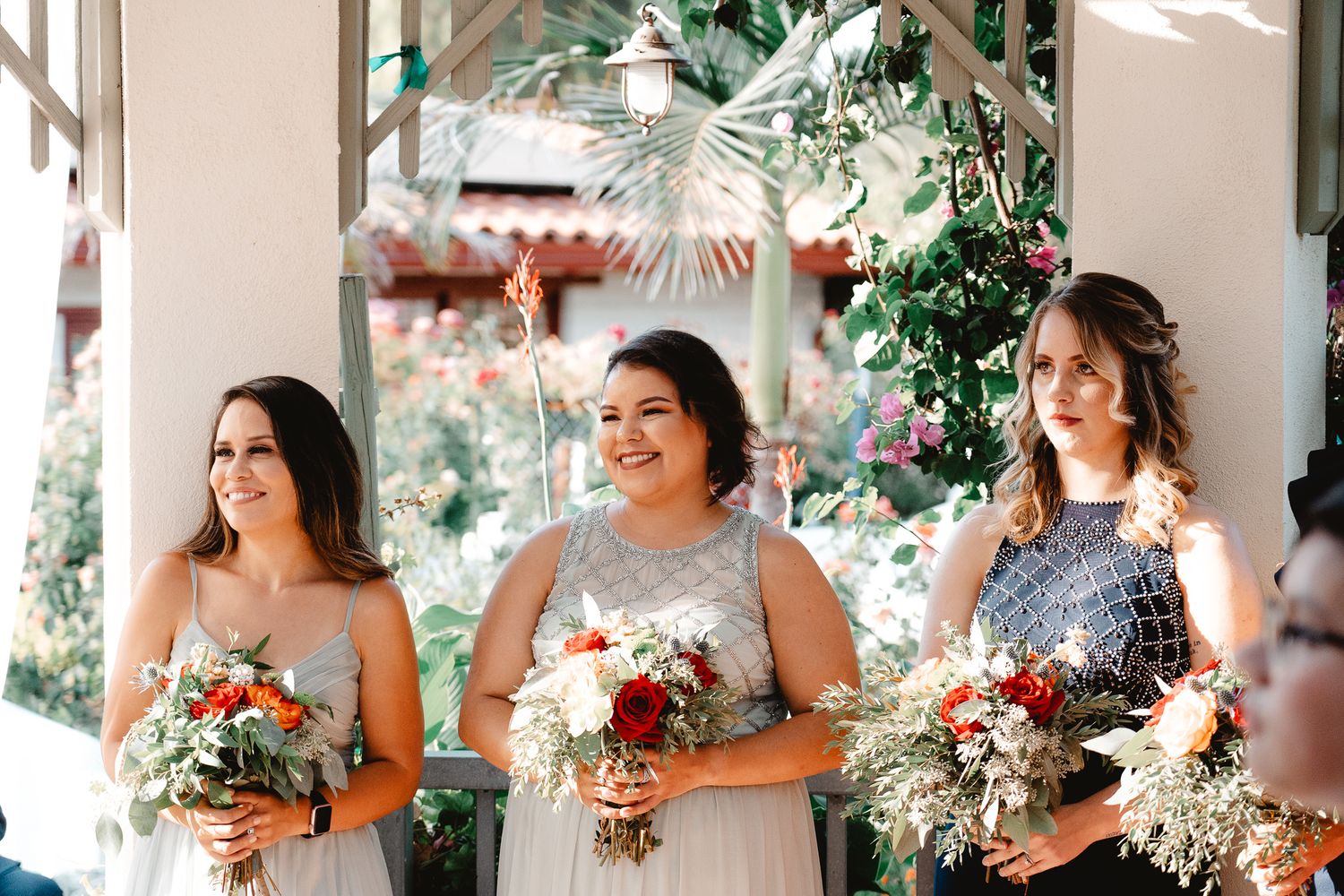 Three bridesmaids in flowing dresses holding colorful bouquets pose together at an elegant outdoor venue.