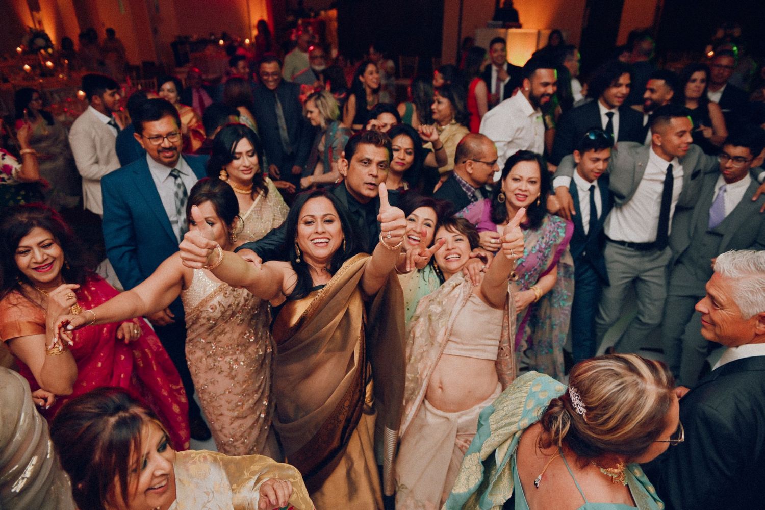 Festive wedding reception with guests dancing and celebrating in formal attire at an indoor venue with warm lighting.
