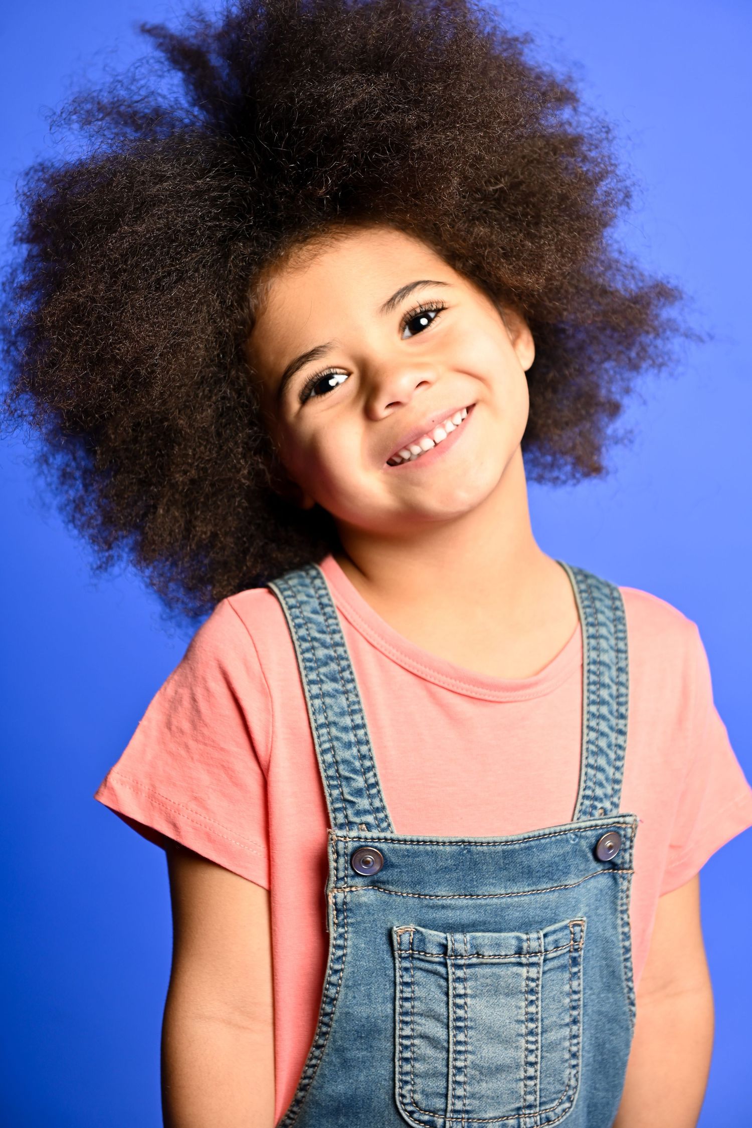 Cheerful child with natural afro hair wearing denim overalls and coral t-shirt against blue background.