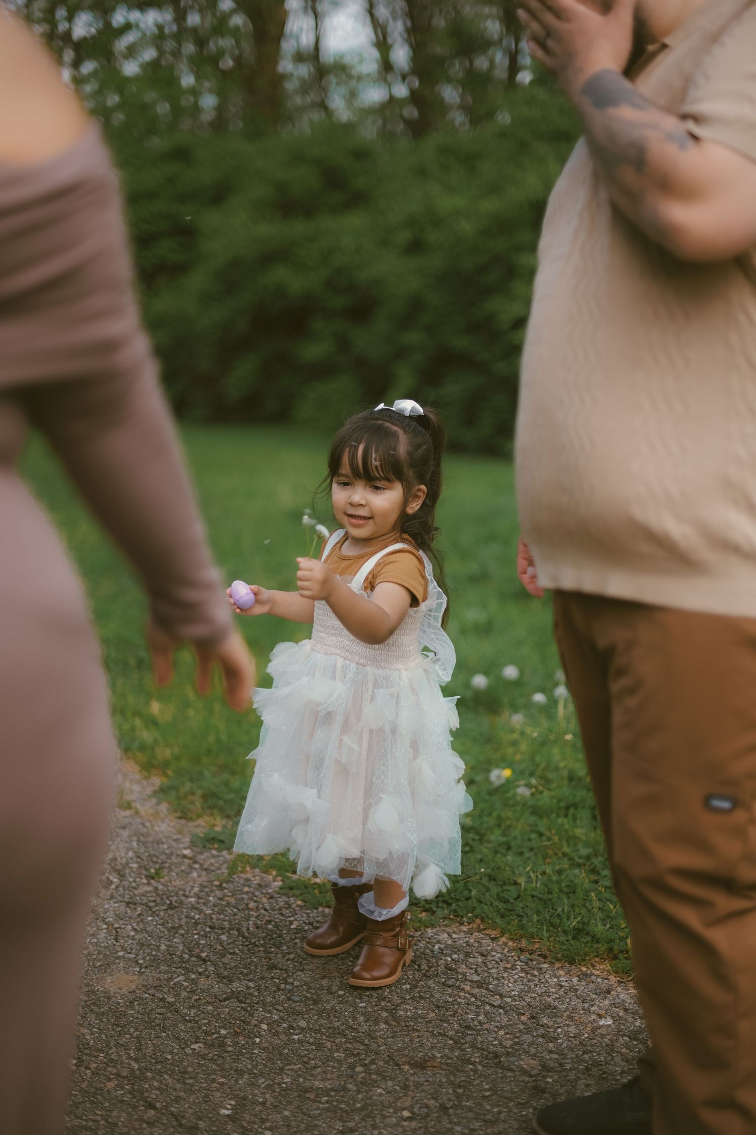 Little girl in white dress holds something while standing between adults on outdoor path.