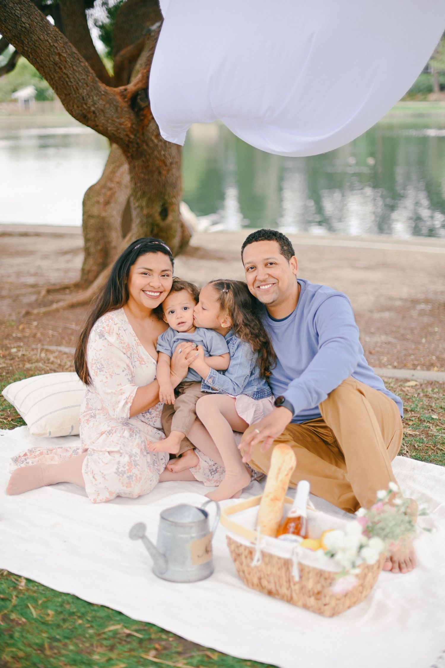 Family enjoying picnic by lake under tree with white fabric and picnic basket.
