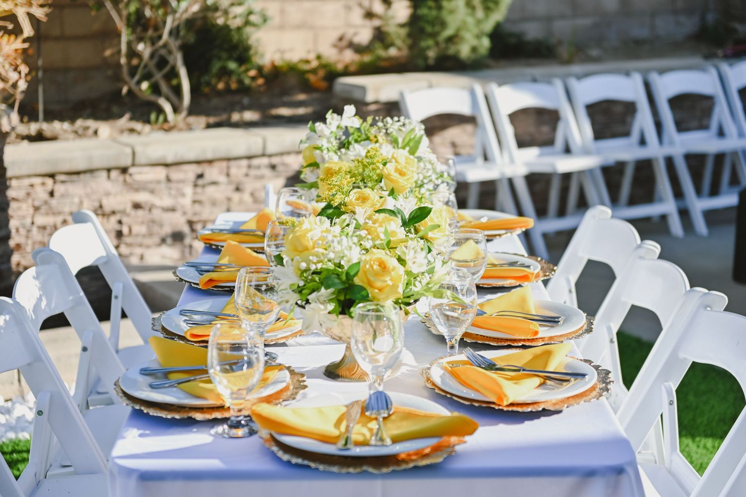 Elegant outdoor wedding table setting with white chairs, yellow napkins and floral centerpiece.