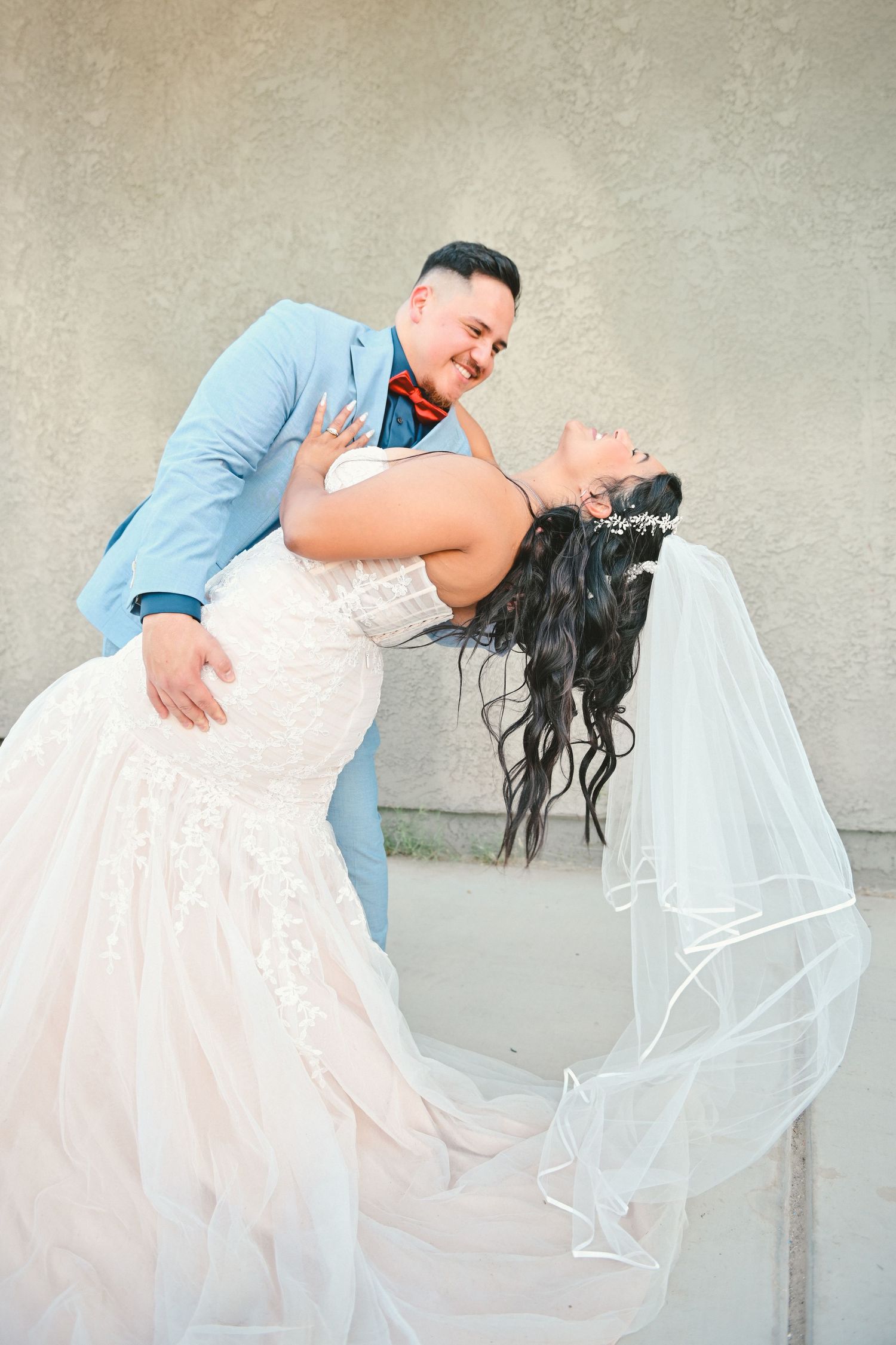 Romantic wedding couple poses with bride dipping back in her lace gown and flowing veil against neutral wall.