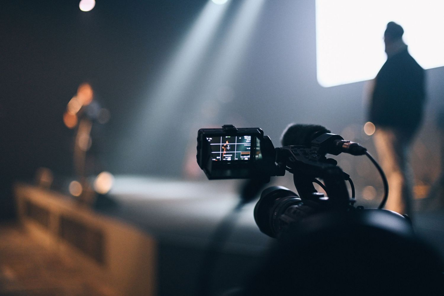 Professional video camera filming an event with stage lighting and silhouettes in the background.