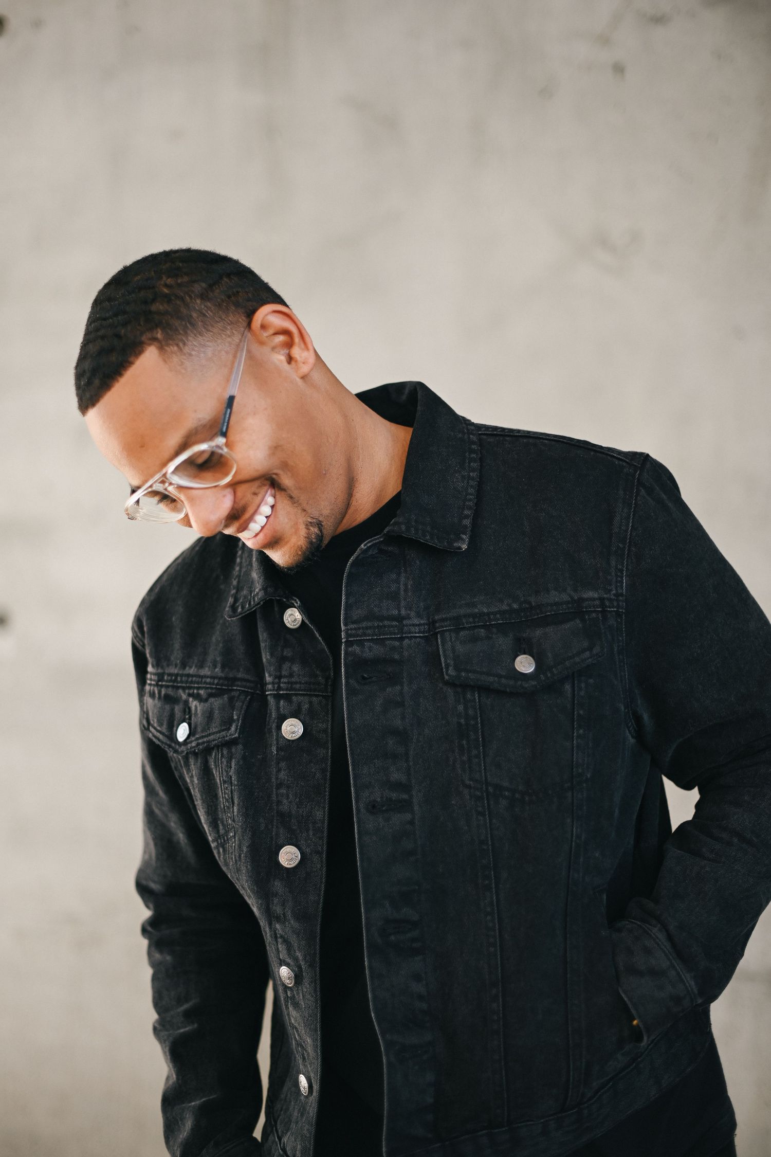 Portrait of person in black denim jacket laughing while looking down against neutral background.