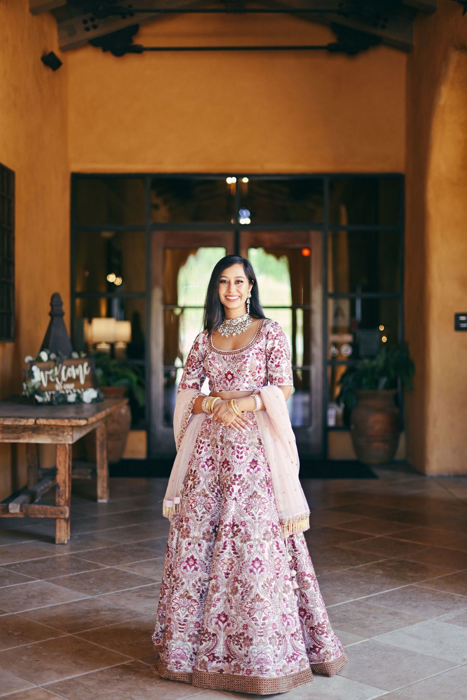 Traditional Indian bridal lehenga in pale pink with intricate red embroidery shown in an elegant indoor setting.