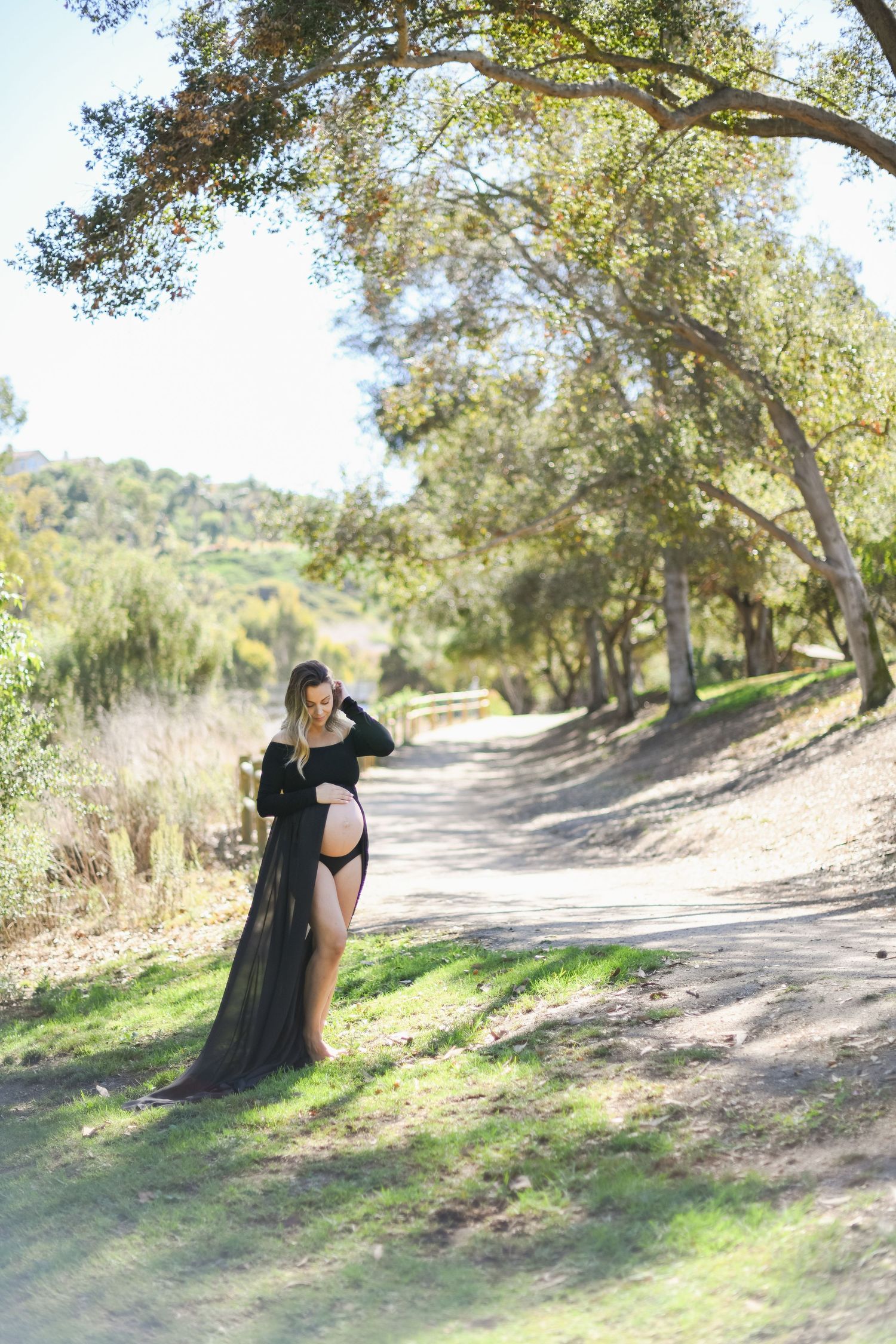 Maternity photo of person in black sheer dress posing on nature trail with sunlit trees.