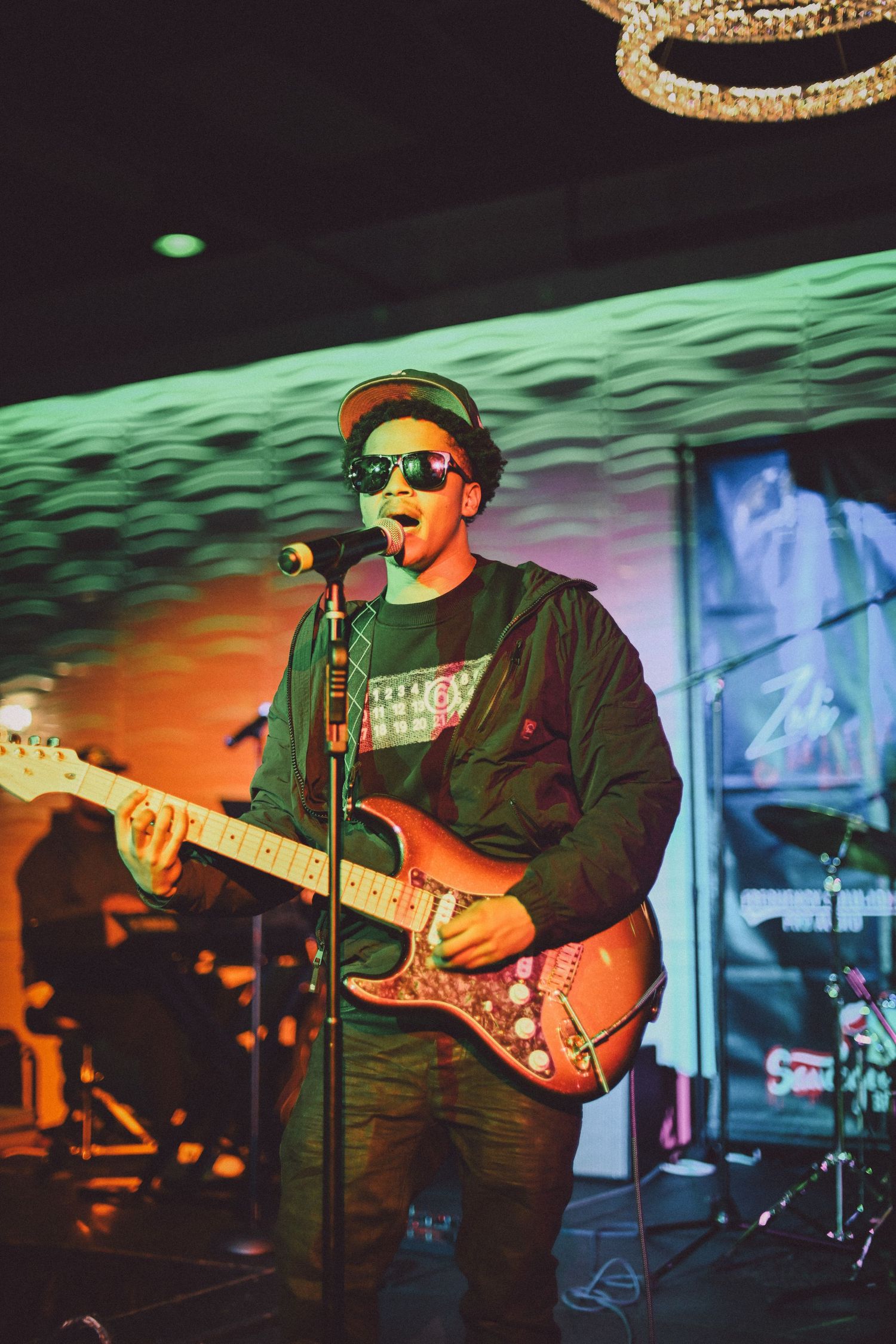 Musician performing with guitar and microphone against colorful stage lighting.