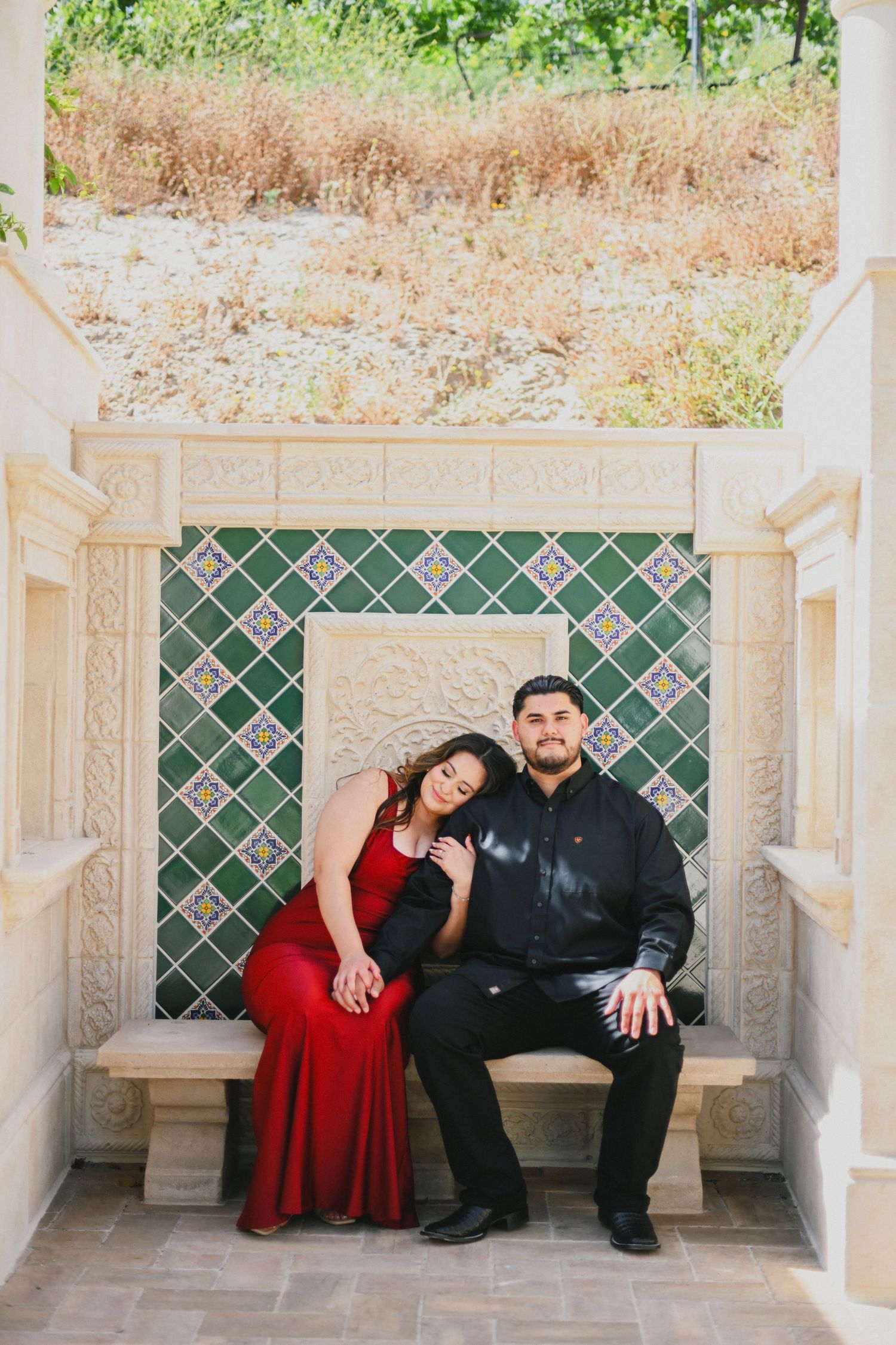Couple in formal attire poses on decorative tiled bench in garden alcove.
