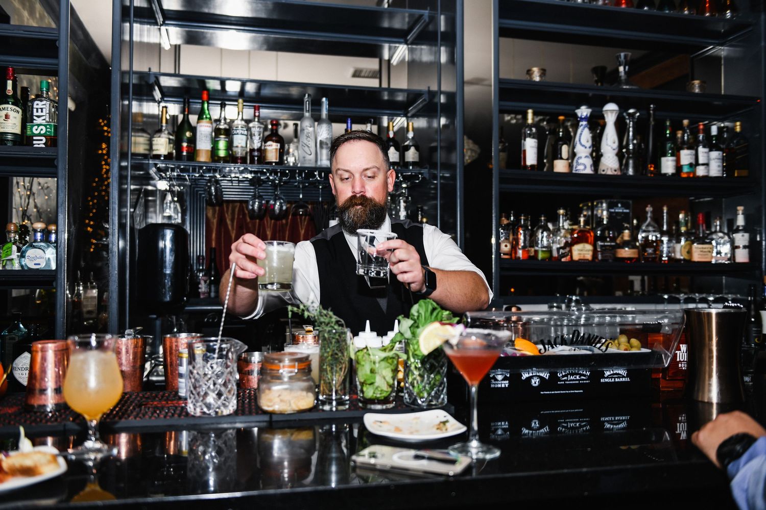 Bartender mixing cocktails behind fully stocked bar with colorful drinks on display.