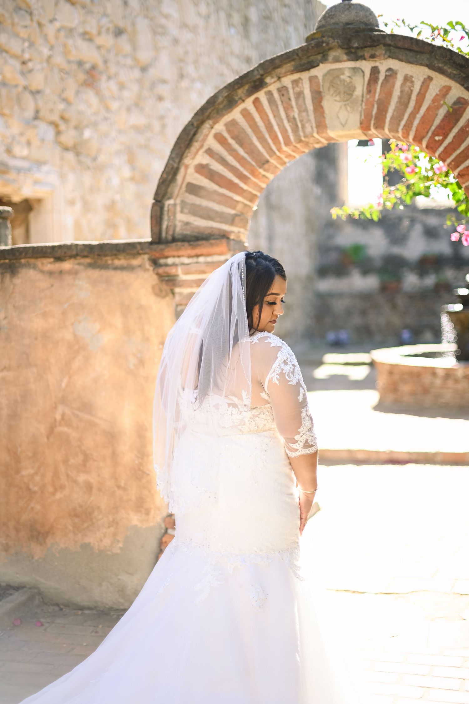 A bride in a lace wedding dress poses in front of a rustic stone archway.