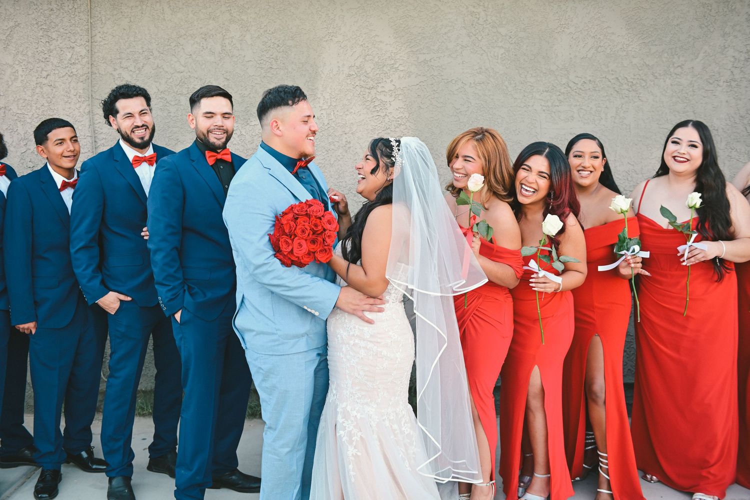 Wedding party wearing navy suits and red bridesmaid dresses pose together against neutral wall with red bouquets.