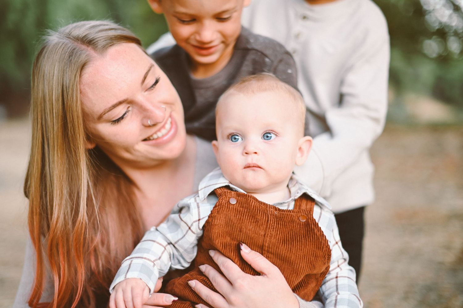 Woman in casual attire smiling while holding baby in brown corduroy outfit outdoors.