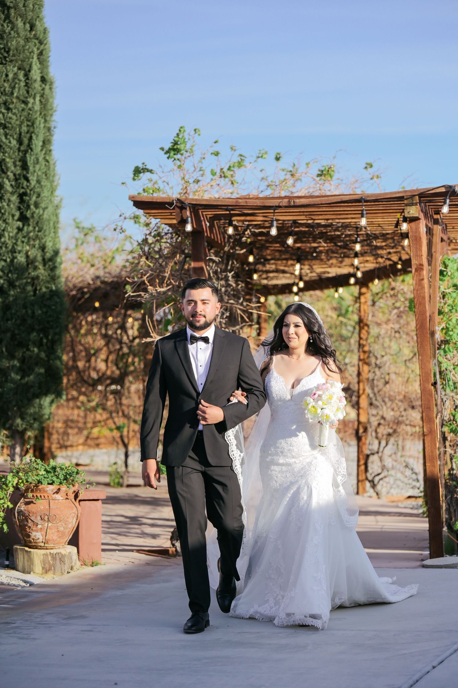 Bride and groom walking under rustic wooden pergola with string lights and greenery.