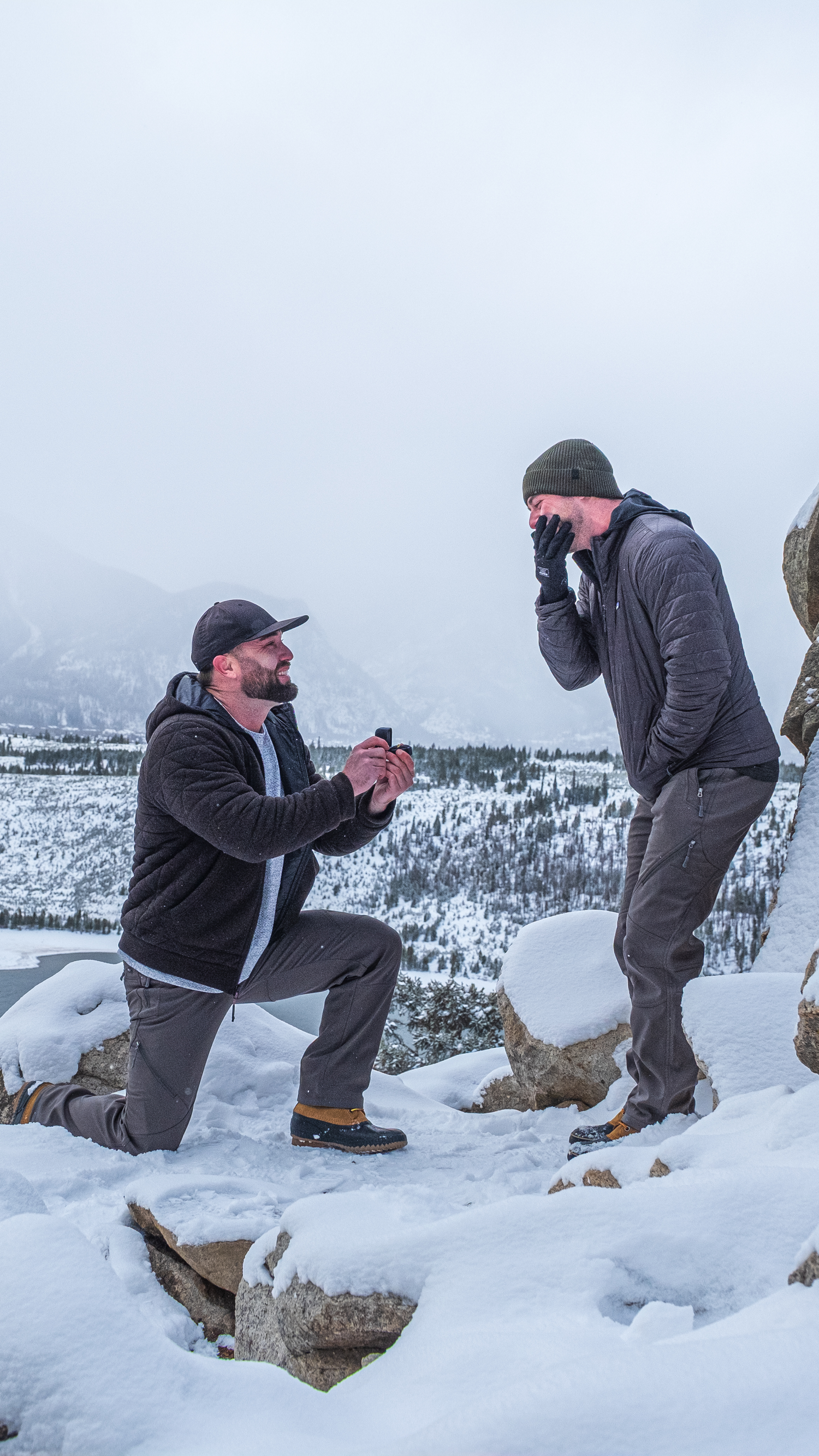 Same sex winter proposal moment captured at snowy Sapphire Point Overlook with snow-covered forest vista in the background.