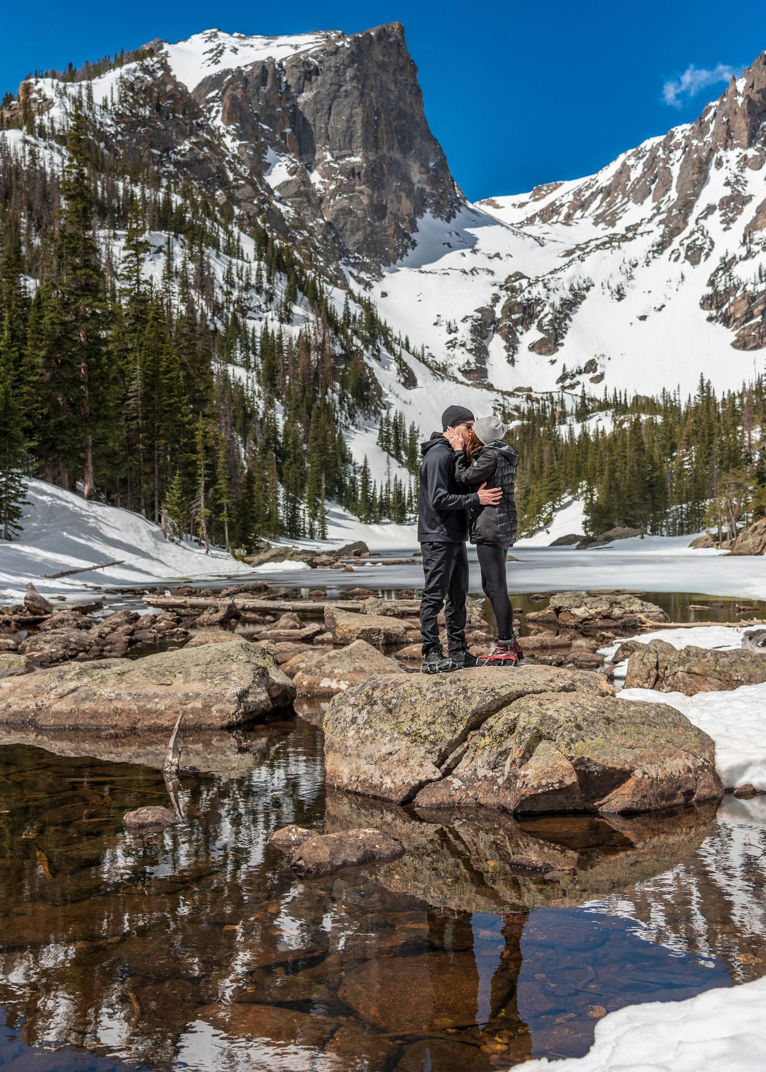 Couple embracing in front of pond inside RMNP Dream Lake after their surprise marriage proposal .