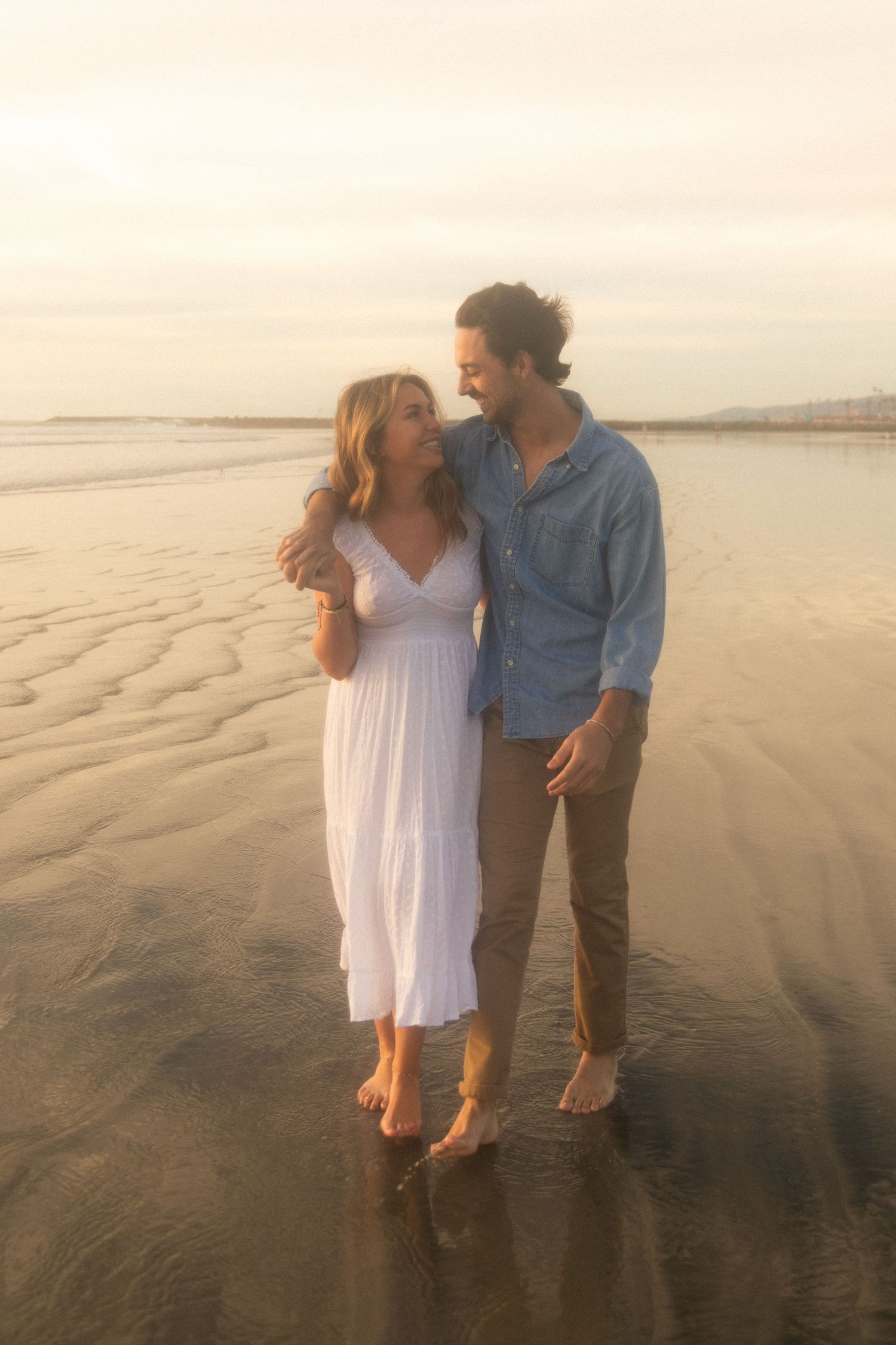 A couple embraces while standing barefoot on a wet beach at sunset, with the ocean waves visible in the background.