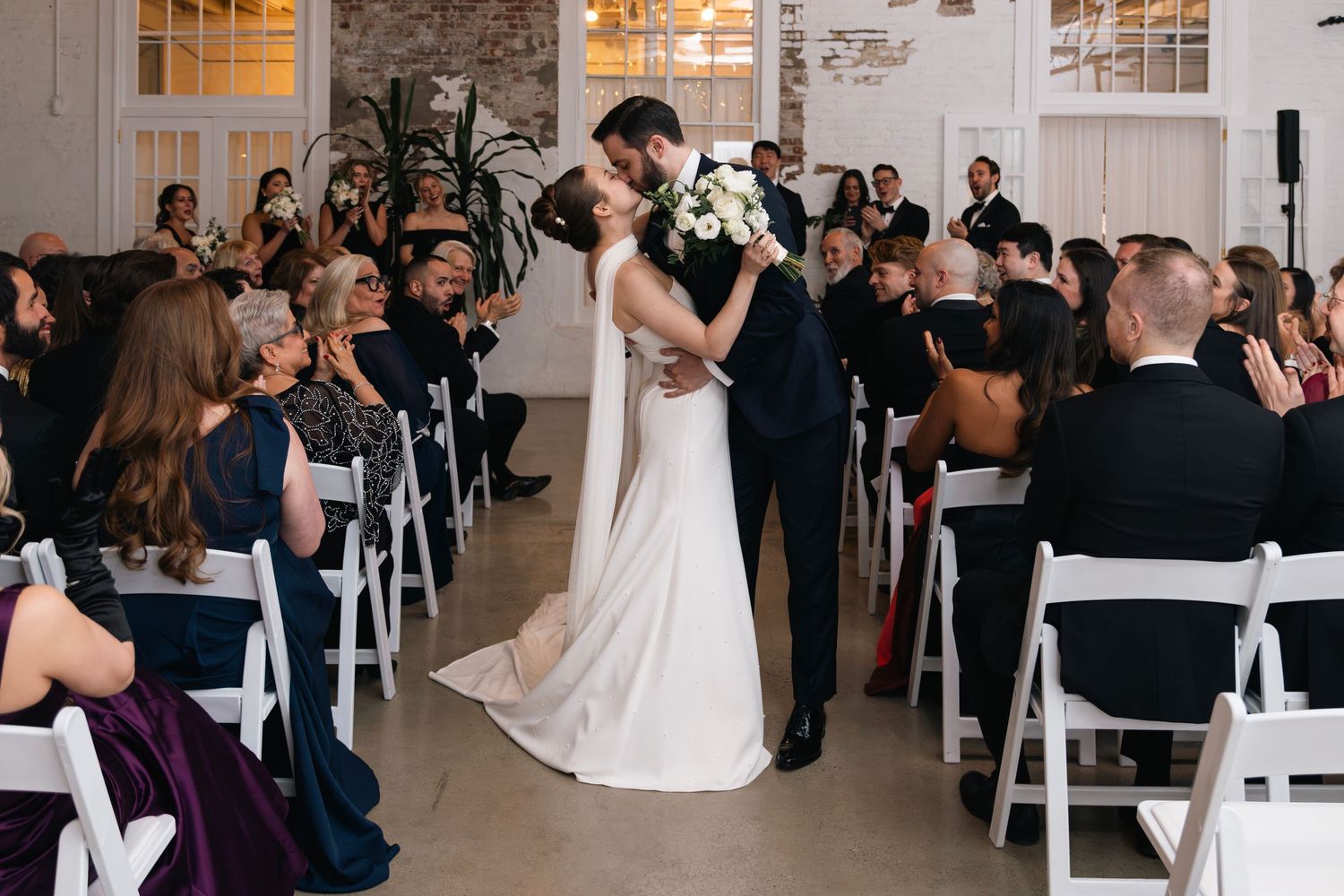 A couple shares their first dance surrounded by wedding guests seated in white folding chairs in an indoor venue.