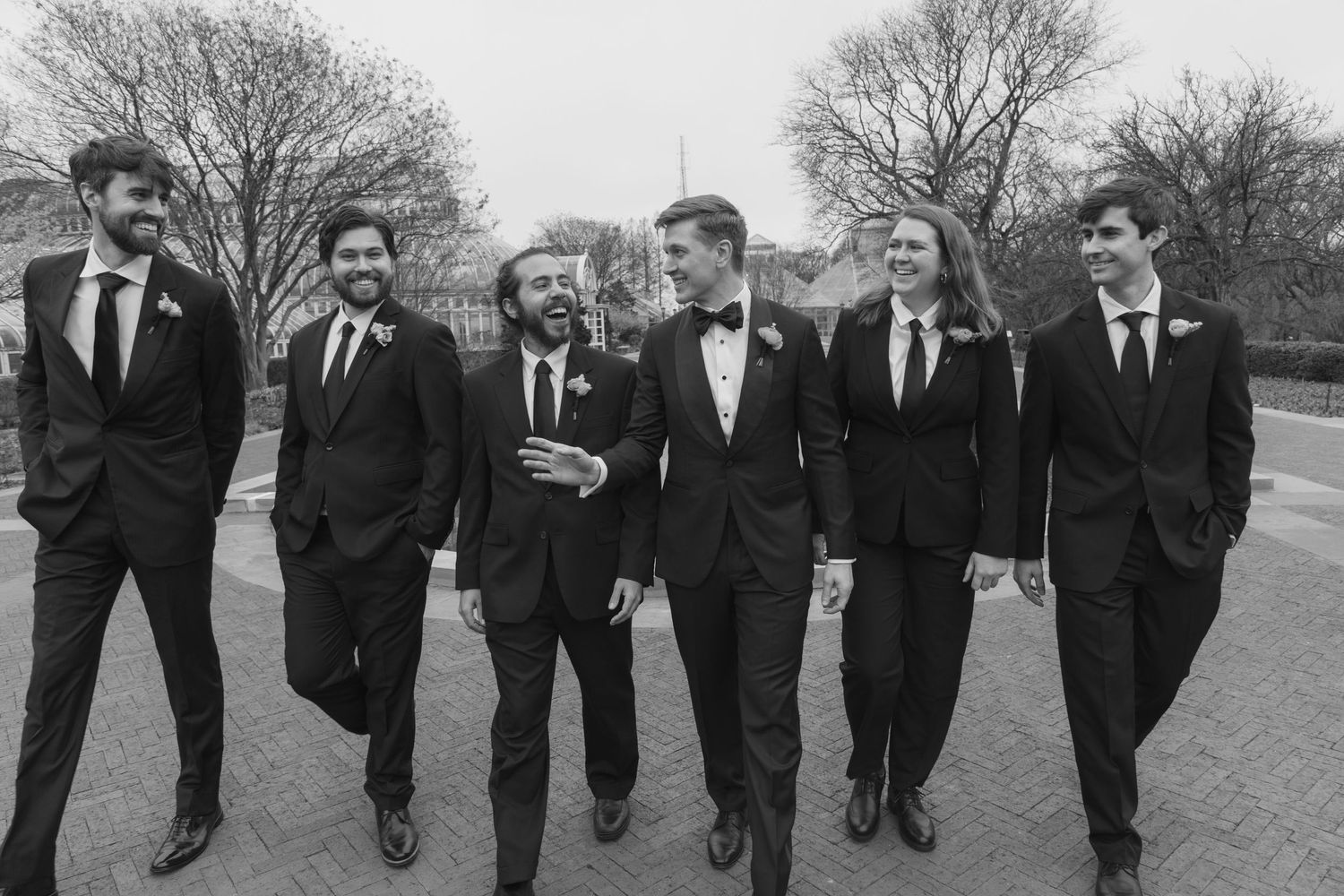 A group of groomsmen in dark suits walk together on a path in black and white.