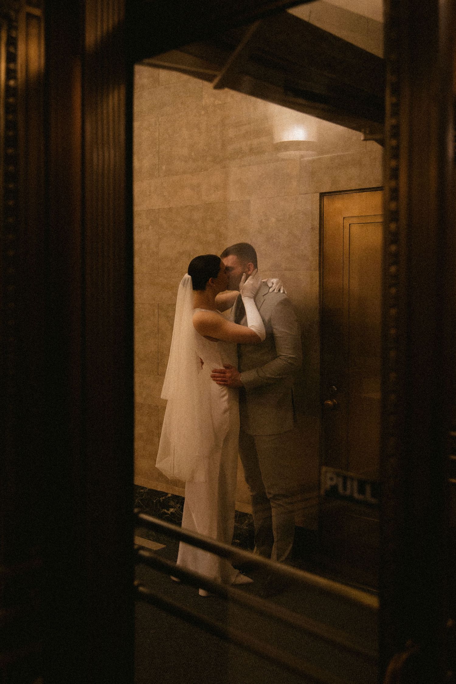 Bride and groom sharing a romantic kiss in a warmly lit elegant doorway, captured through wooden frames.