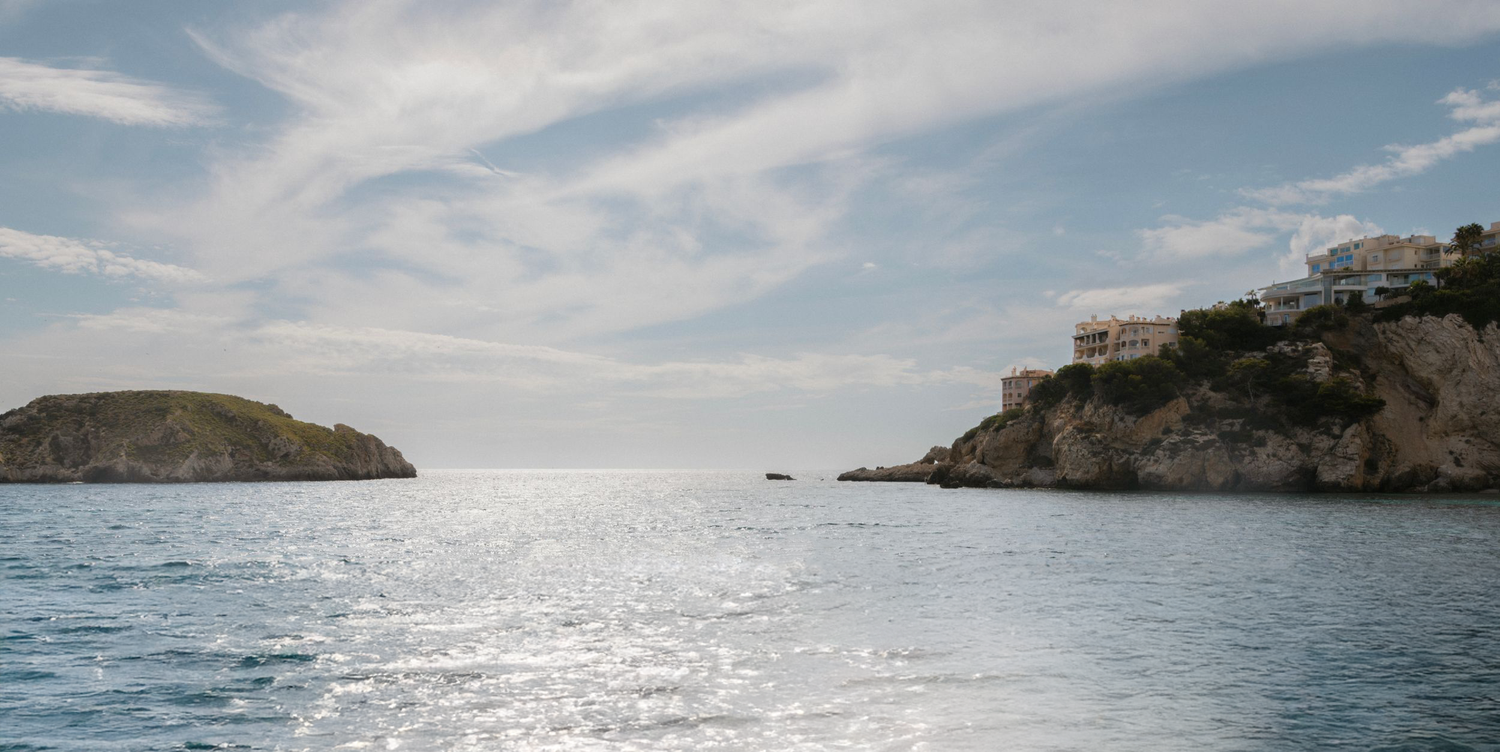 A scenic Mediterranean coastal view with sunlit cliffs and islands rising from the sparkling sea under wispy clouds.