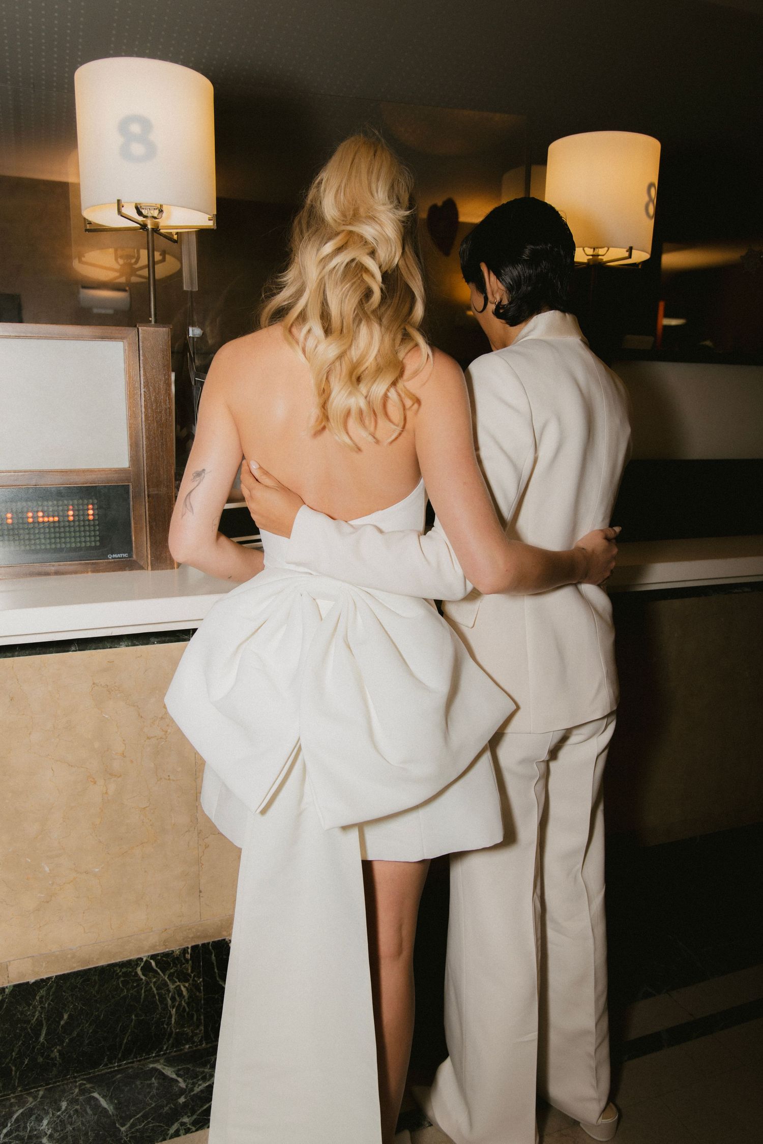 A couple in elegant white attire lean against a dimly lit bar counter at an evening event.