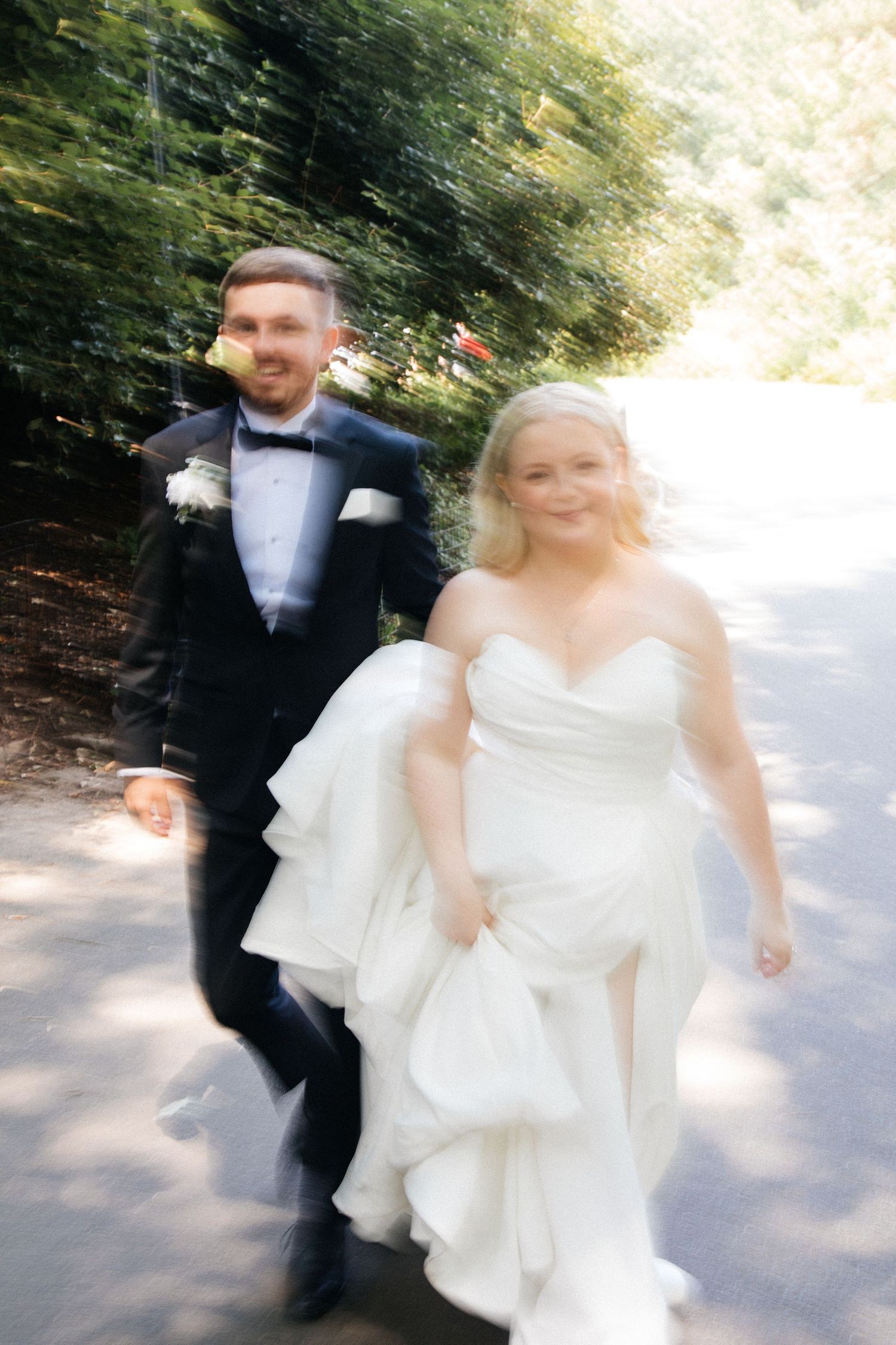 A couple in formal wedding attire walking together on a wooded path, with motion blur creating a dreamy effect.