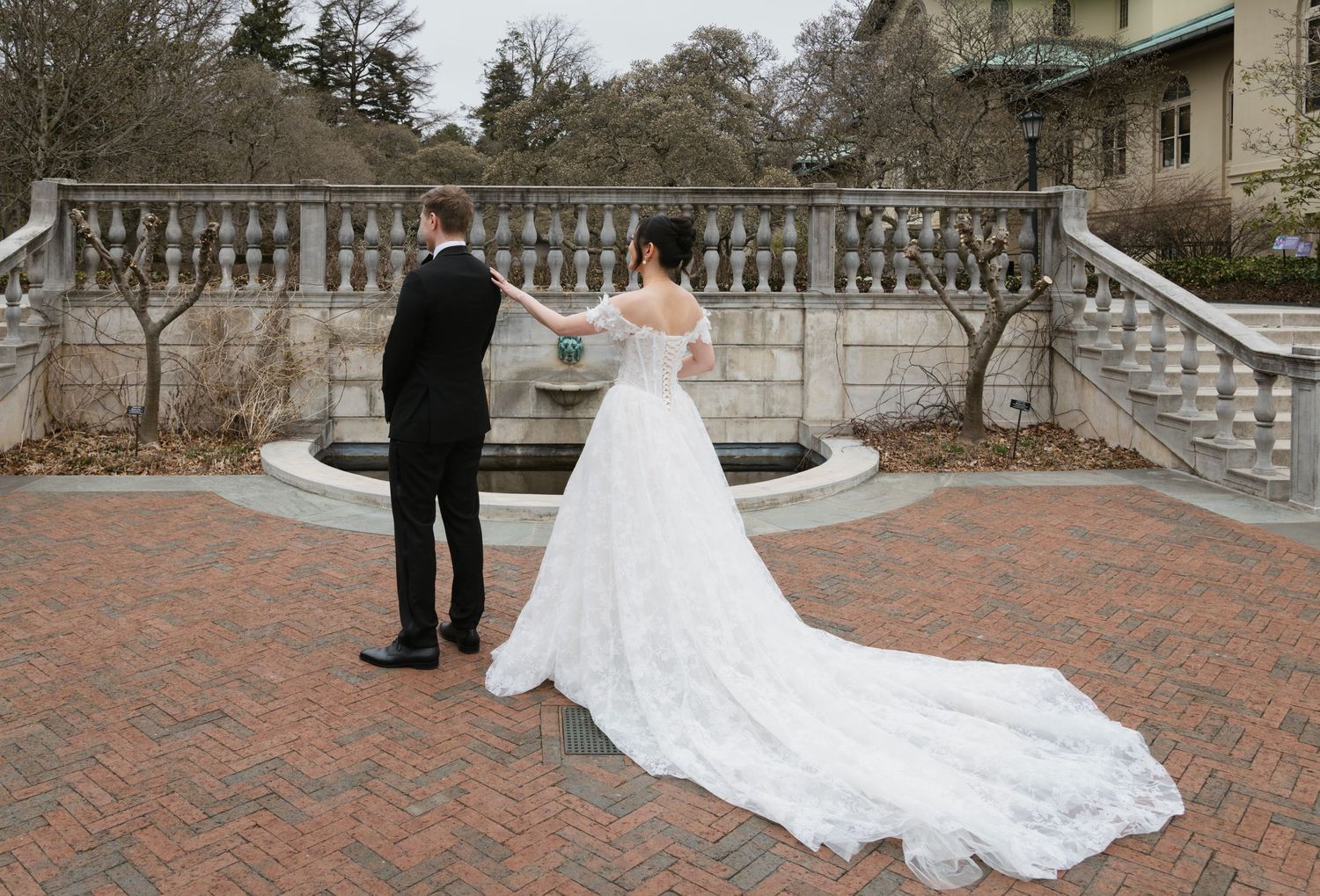 A couple stands on a brick courtyard with an elegant white wedding dress featuring a long flowing train against stone steps.