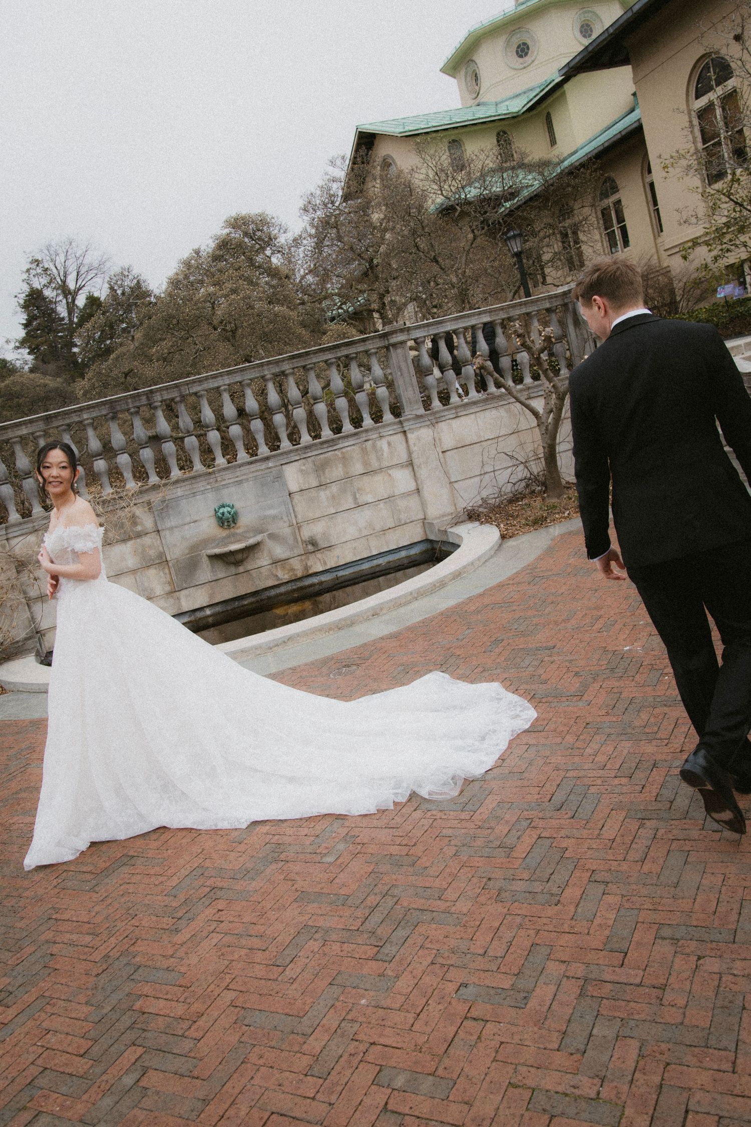 A couple in wedding attire stands on a brick terrace with a beautiful mansion and stone balustrade in the background.