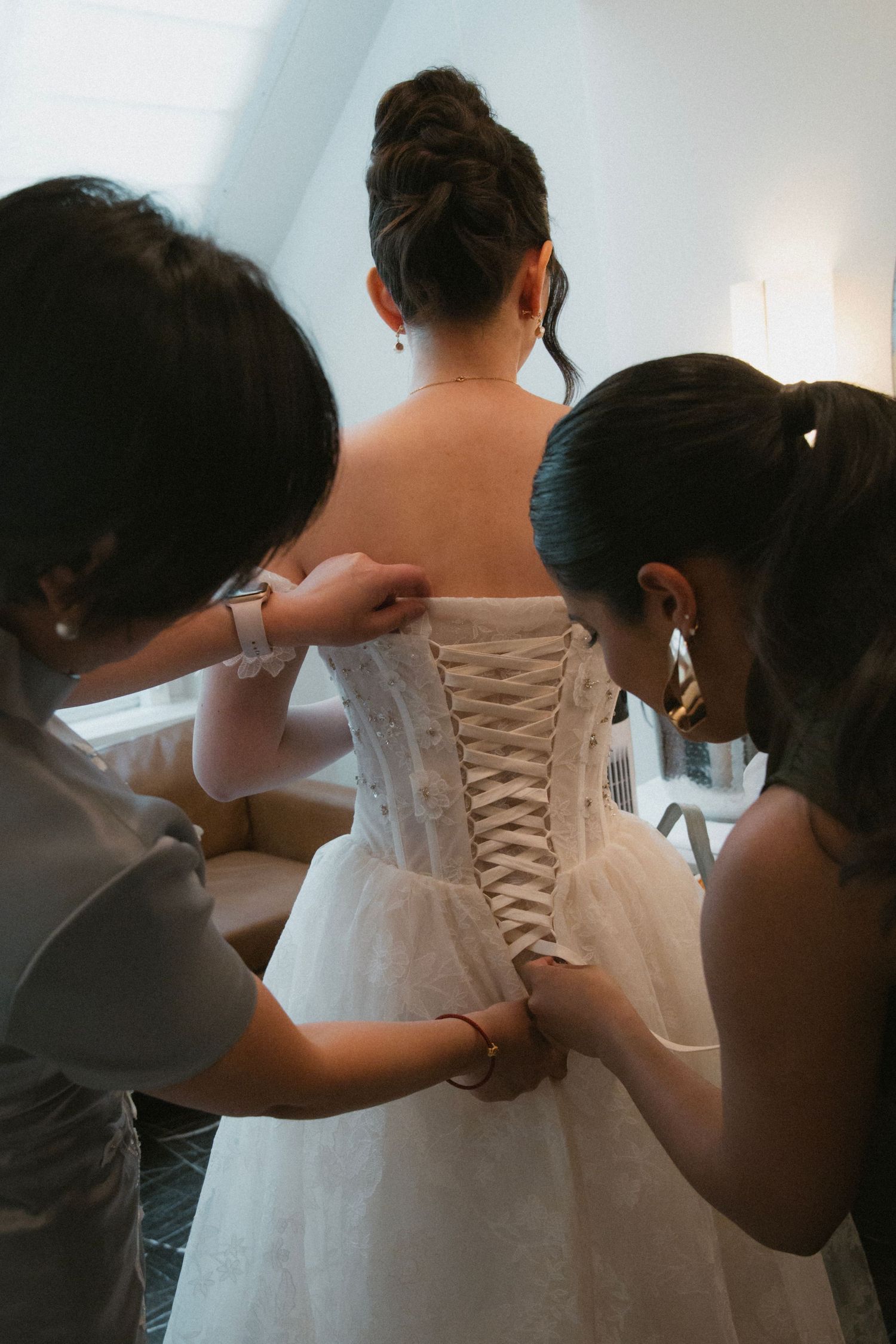 Bridesmaids help lace up the corset back of a white wedding dress before the ceremony.