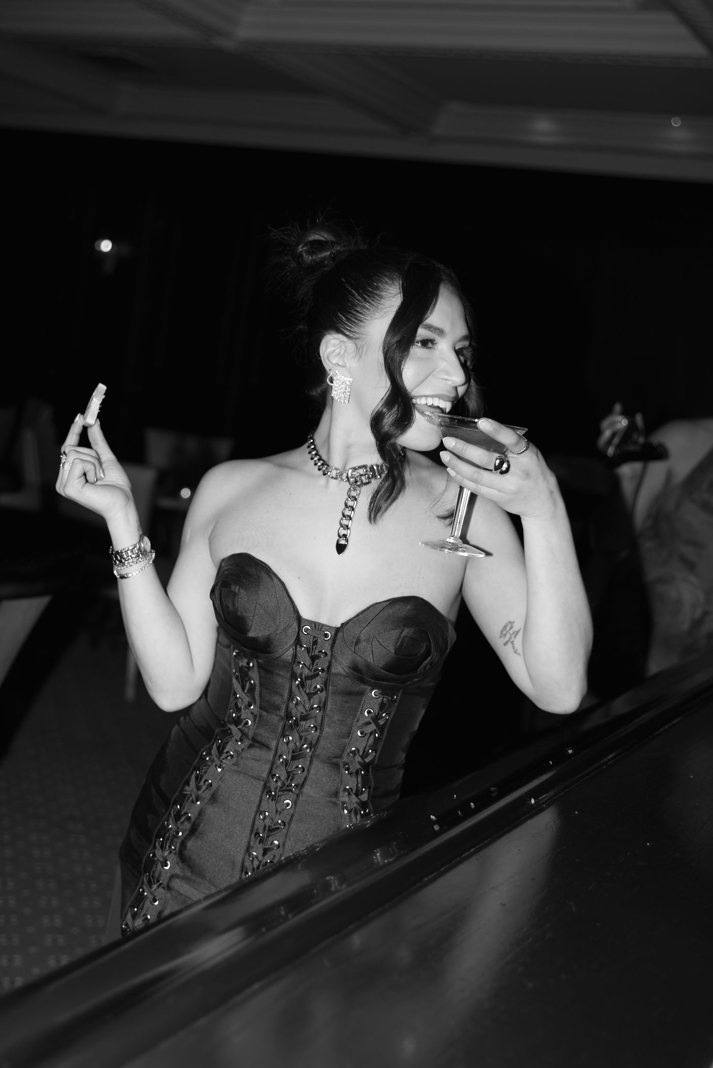 Woman in dark corset-style top seen through window at night with martini glass and moody lighting.