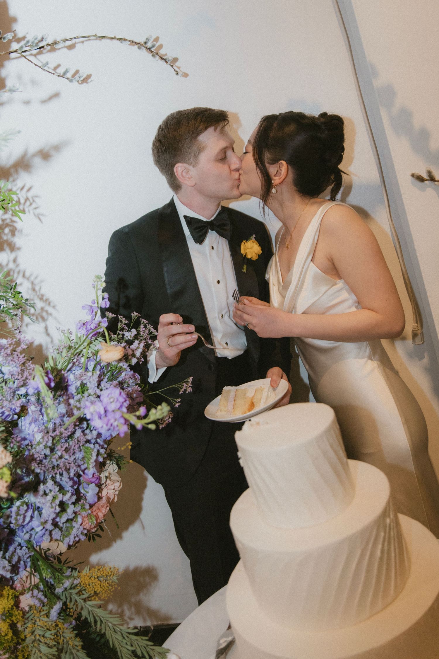 A romantic wedding moment featuring a couple kissing by their tiered white cake, with purple flowers in the decor.
