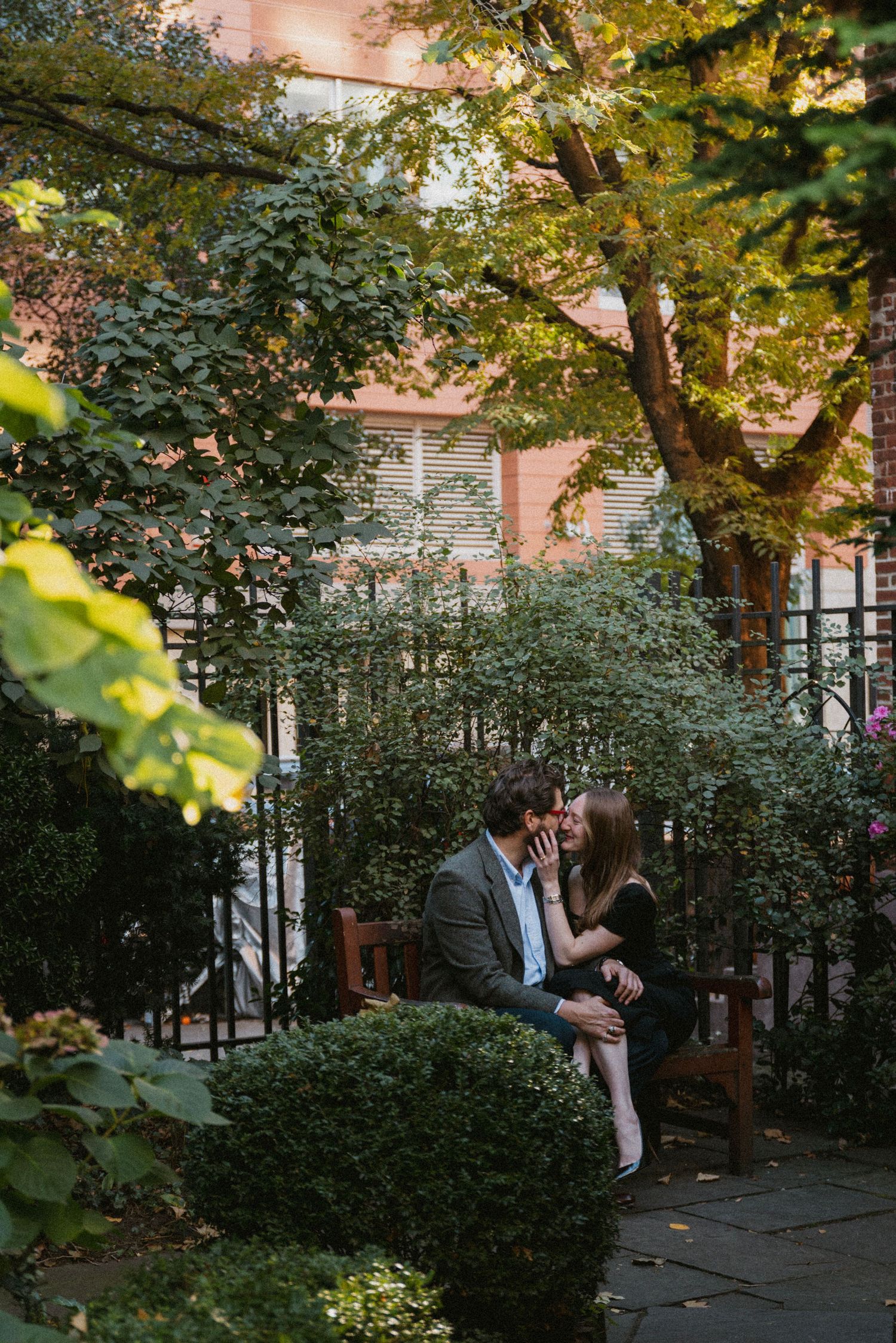A couple shares an intimate moment among lush greenery and trees in an urban garden setting at sunset.